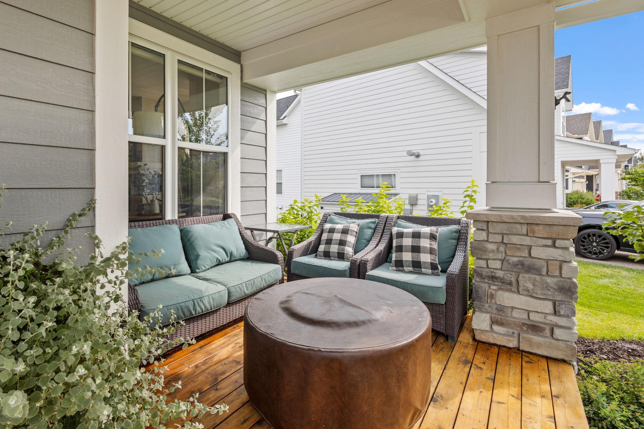 Covered porch with wicker seating, teal cushions, plaid pillows, and a round ottoman on wood decking.
