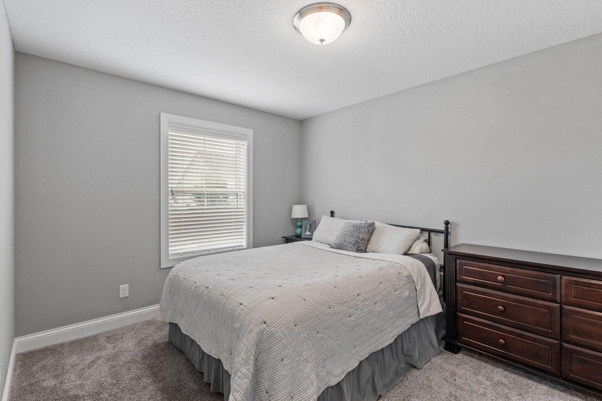 Bedroom with a large gray-covered bed, carpet, window blinds, and dark wooden dresser.