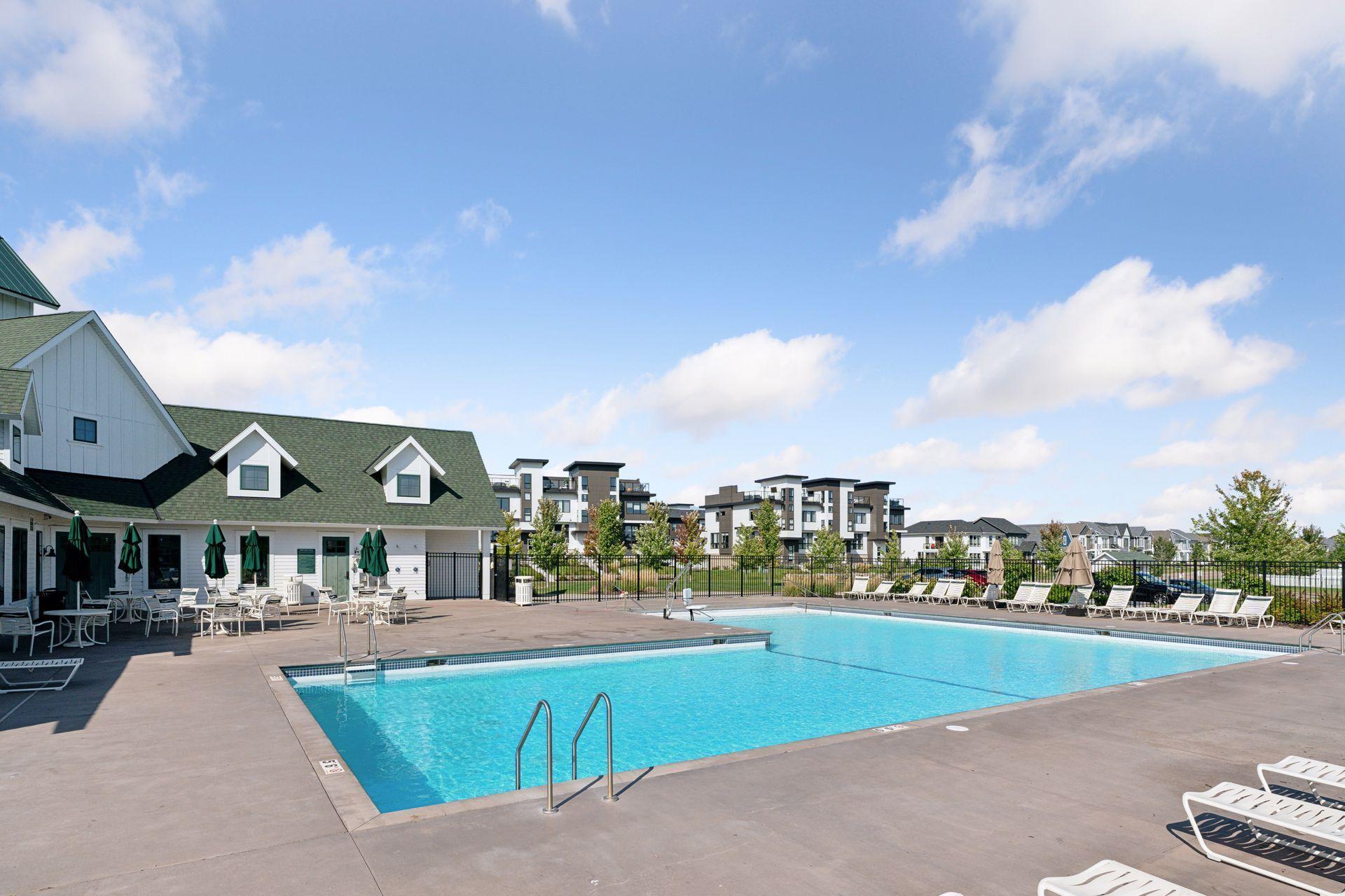 Community pool with lounge chairs and umbrellas beside a clubhouse and apartment buildings.