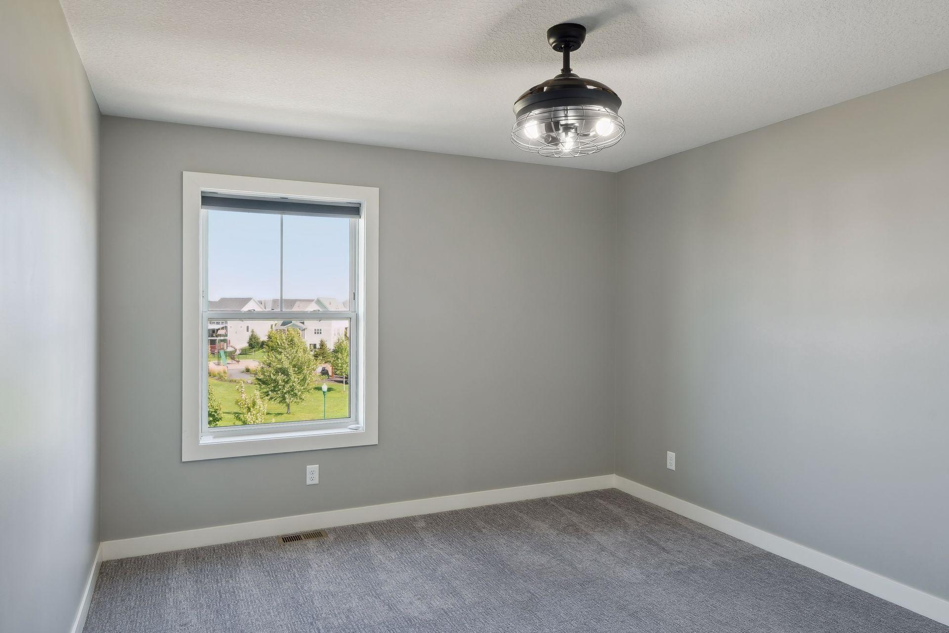 Empty gray room with carpet, a ceiling fan, and a window overlooking trees and a playground.