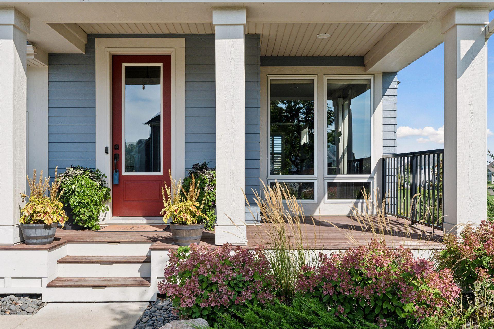 Blue-sided home’s covered porch with a red front door, white columns, and flowering plants.