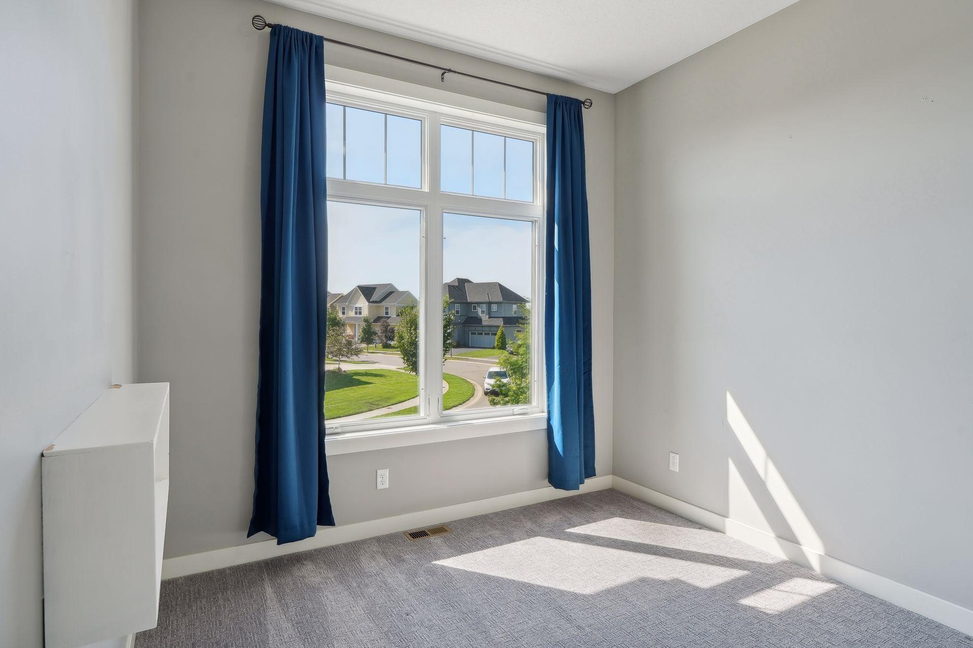 Vacant carpeted room with blue curtains framing a large window overlooking neighboring homes.