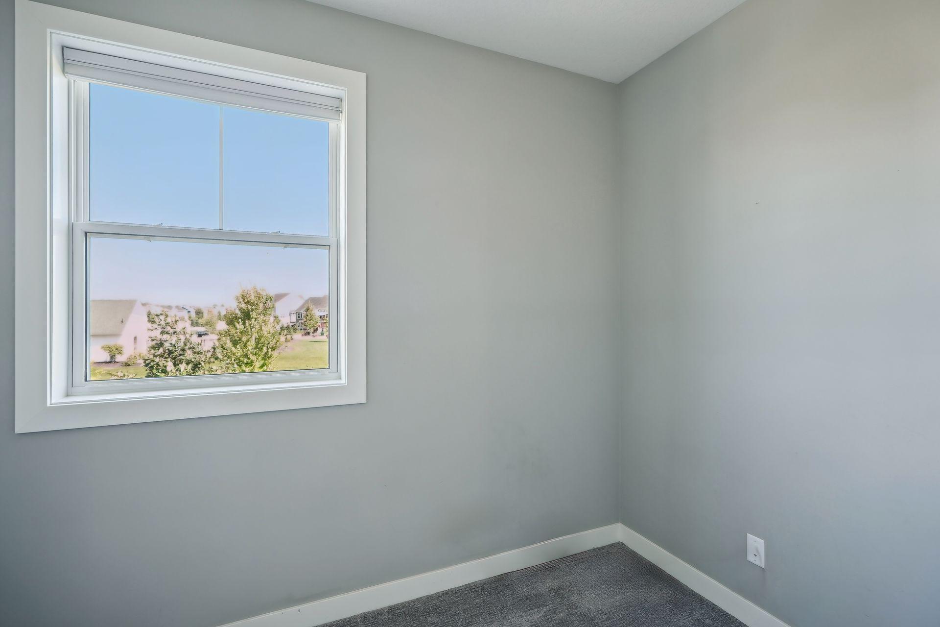 Empty gray-carpeted room with a large window overlooking neighboring homes.