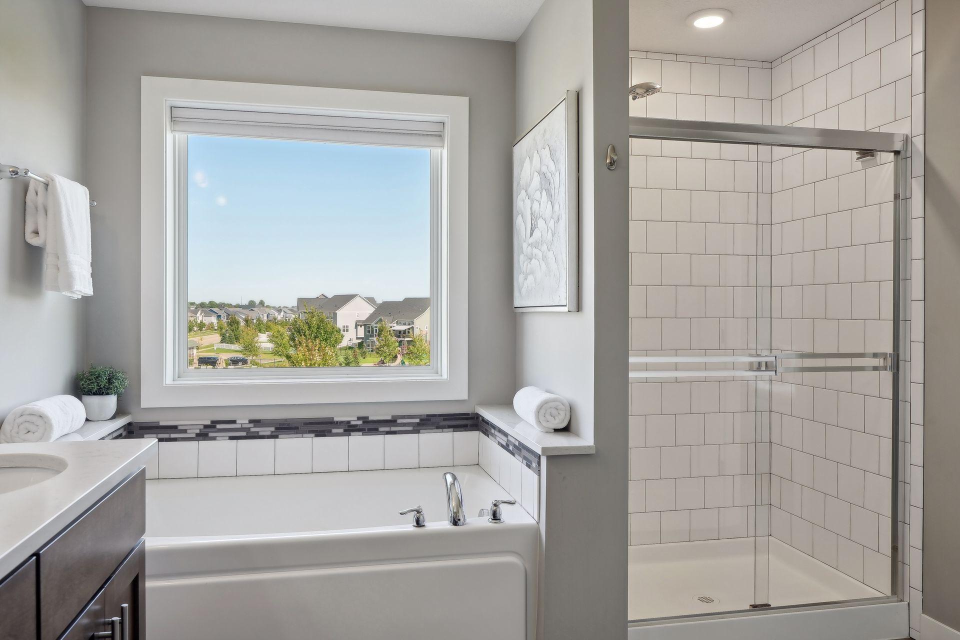 Bathroom with a white tub beneath a large neighborhood-view window and a glass-enclosed tiled shower.
