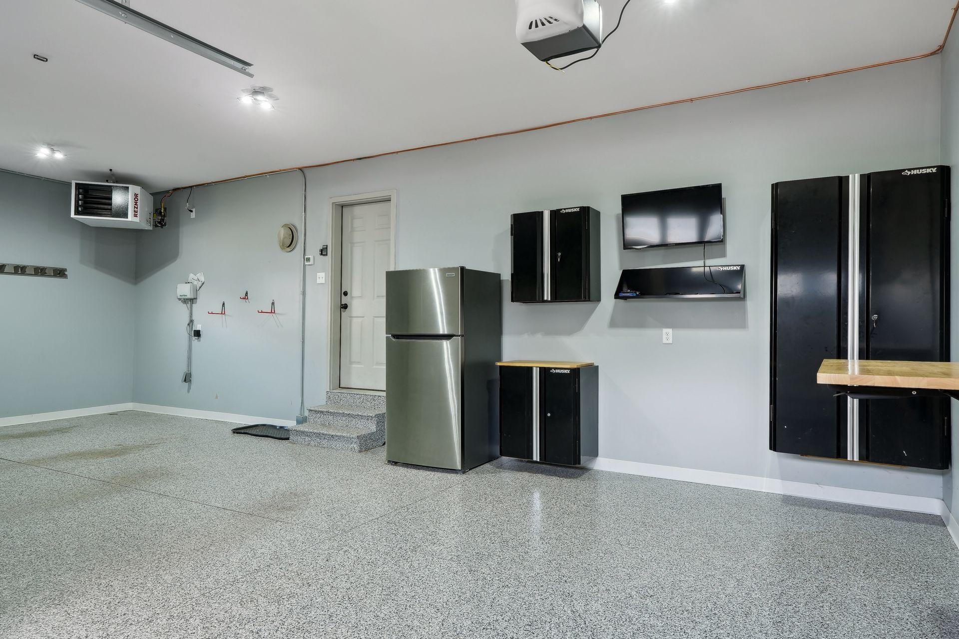 Garage with a speckled floor, black storage cabinets, a refrigerator, and a wall-mounted television.