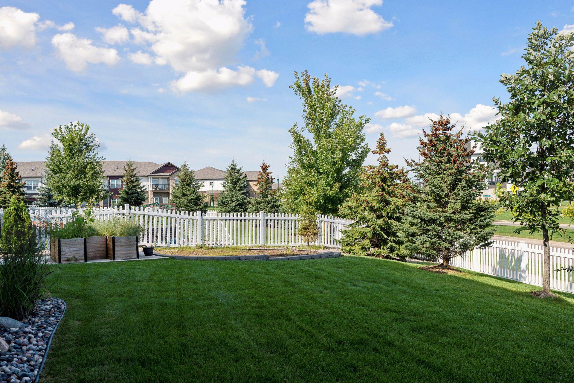 Landscaped backyard with a broad lawn, white picket fence, raised planters, and mature trees.