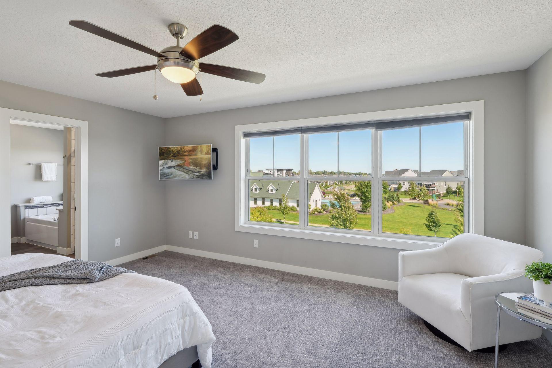 Primary bedroom with a large neighborhood-view window, ceiling fan, and open doorway to the ensuite bathroom.