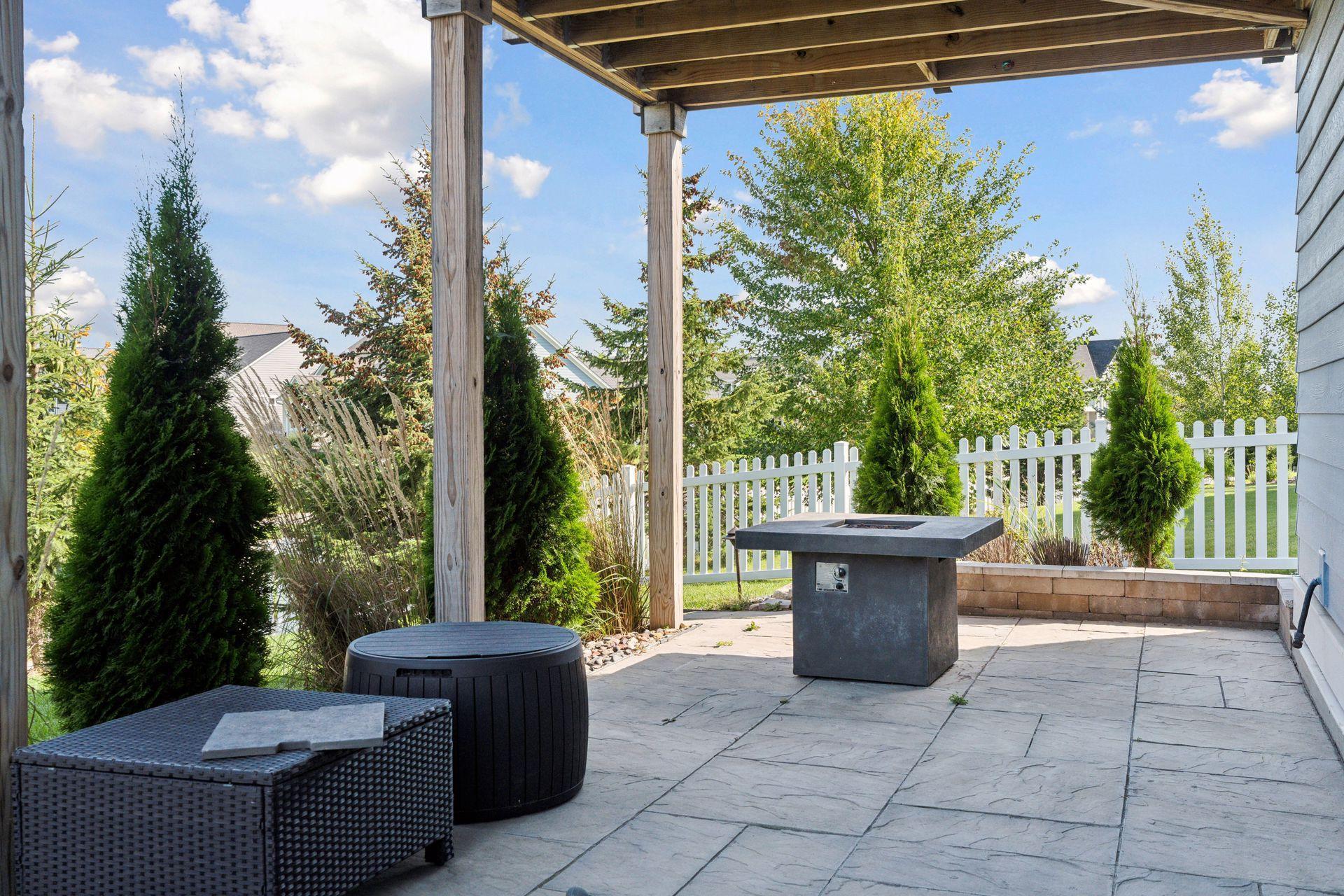 Paved patio beneath a wooden pergola with a gas fire table, woven seating, and landscaped fenced yard.