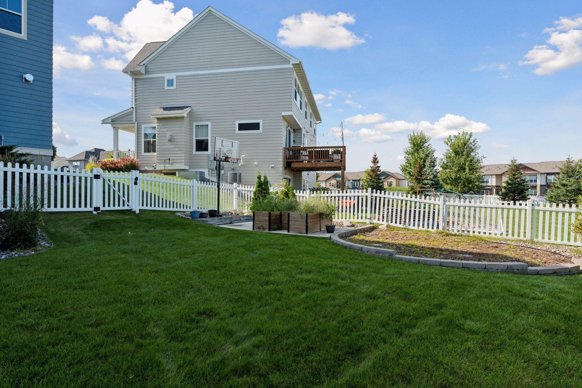 Two-story gray home with a raised deck, fenced garden beds, and a broad green lawn.