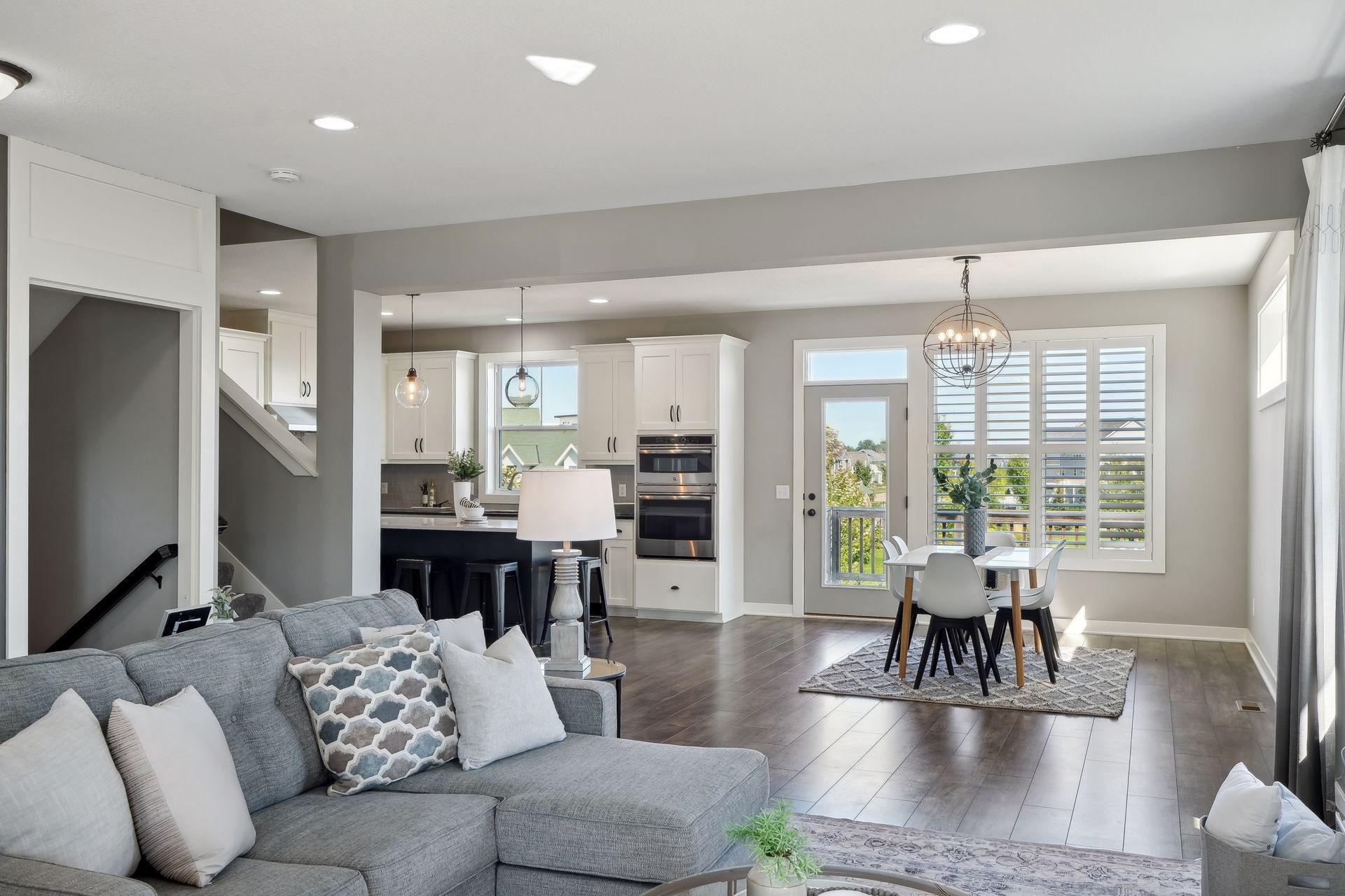 Open-plan living room with gray sectional, white kitchen, and dining area by shuttered windows.