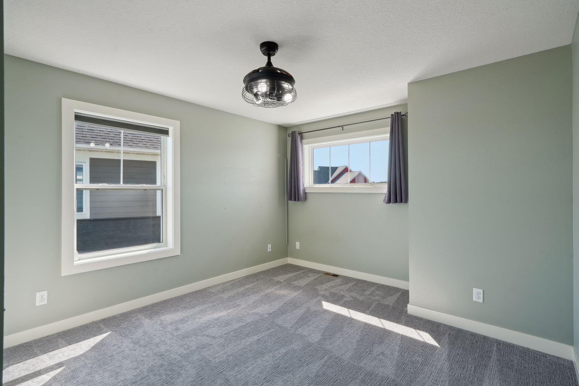 Empty bedroom with sage-green walls, gray carpet, two windows, and a dark ceiling fan.