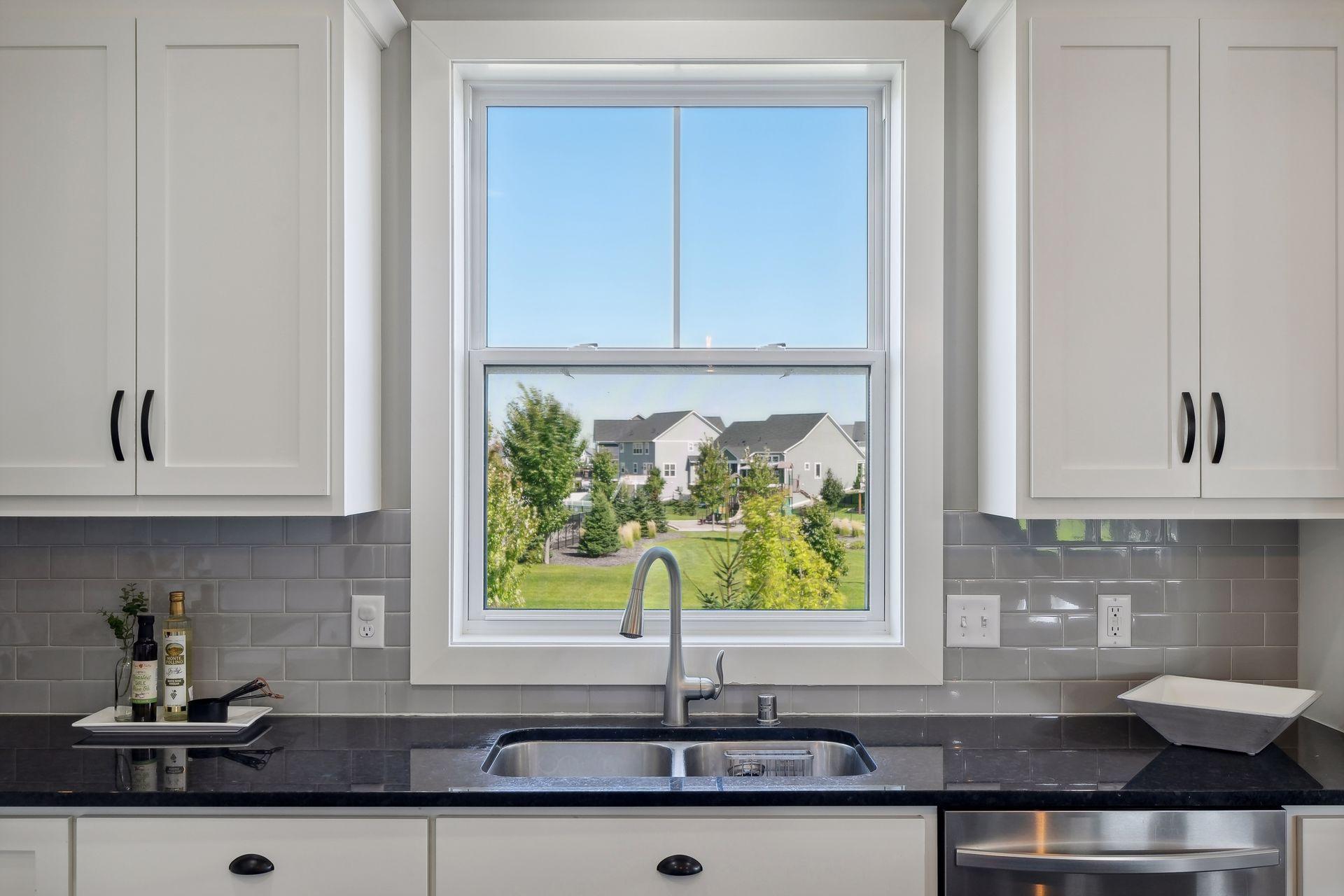 Kitchen with white cabinets and gray tile backsplash, featuring a large window overlooking landscaped grounds and nearby homes.