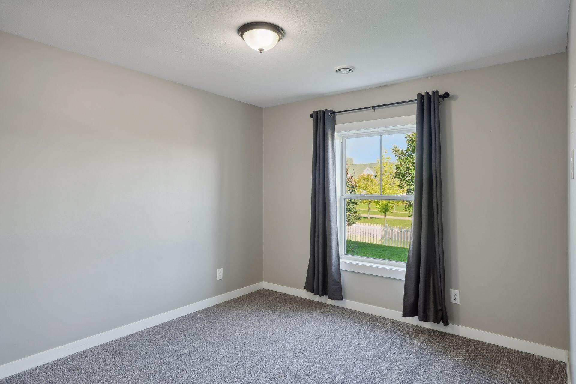 Empty bedroom with gray carpet and dark curtains framing a window overlooking a white picket fence.