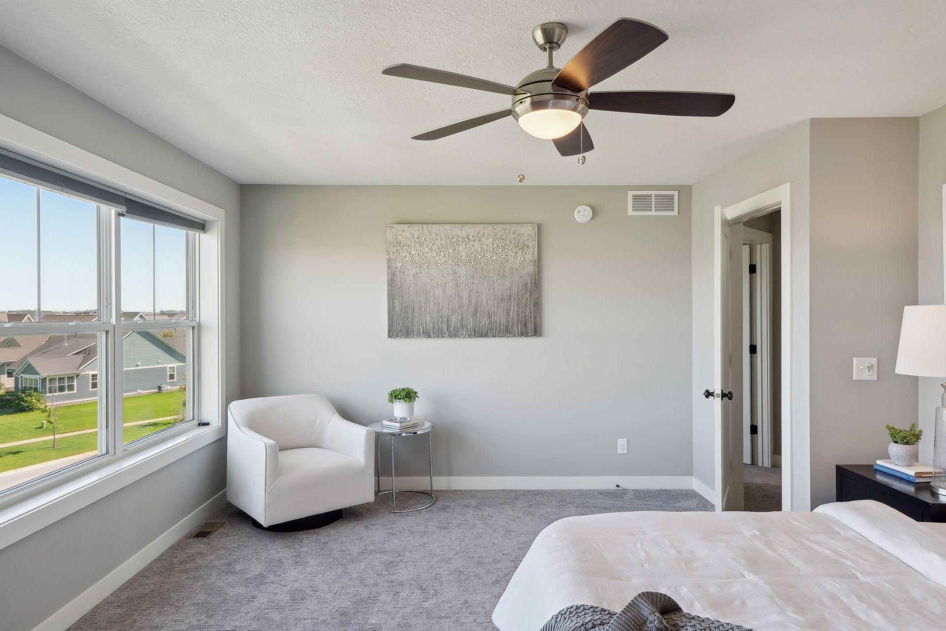 Staged bedroom with a large neighborhood-view window, ceiling fan, and white armchair.
