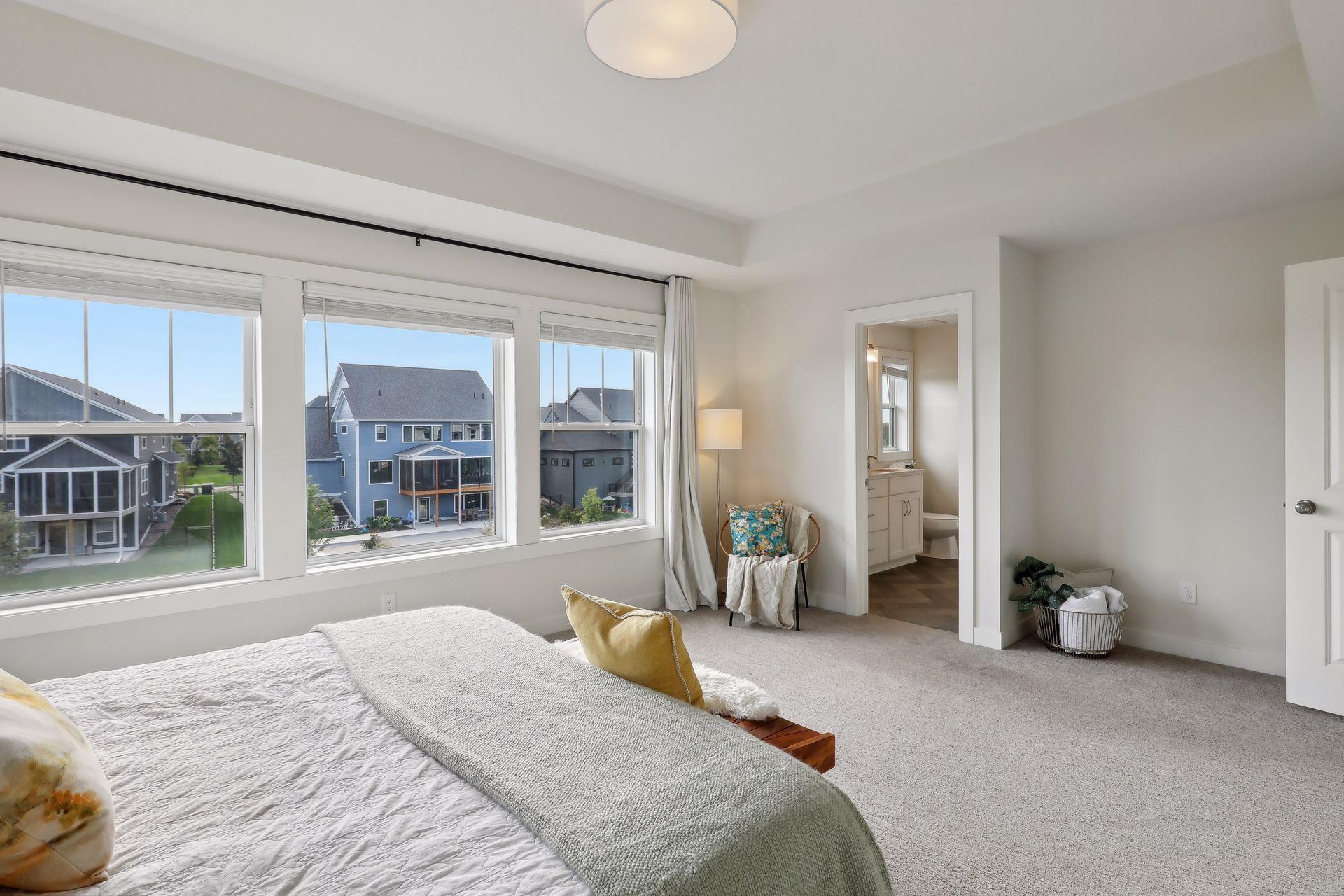 Staged bedroom with three large windows overlooking neighboring homes and an open ensuite bathroom doorway.
