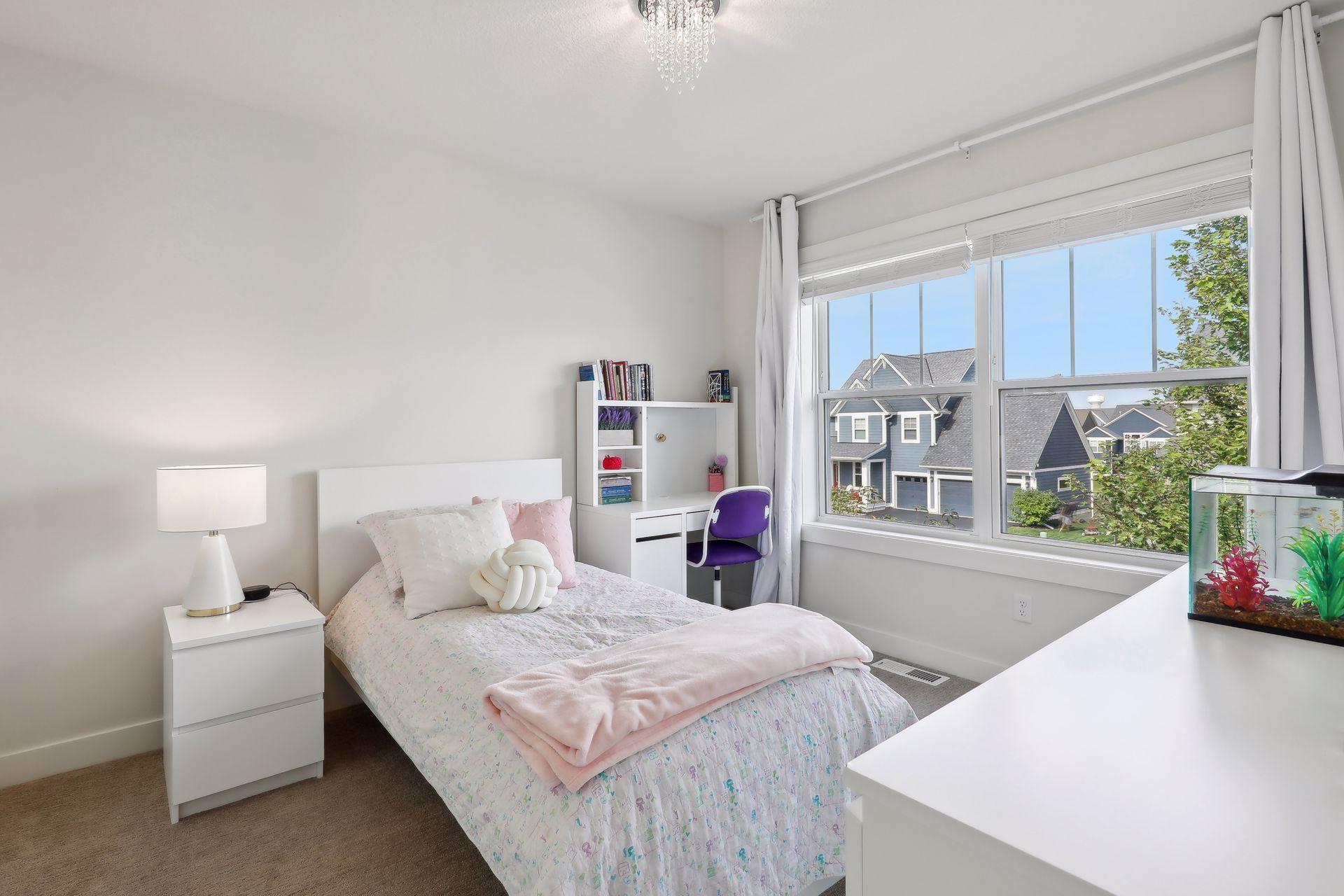 Bright bedroom with pale pink bedding, a purple desk chair, and a large window overlooking neighboring homes.