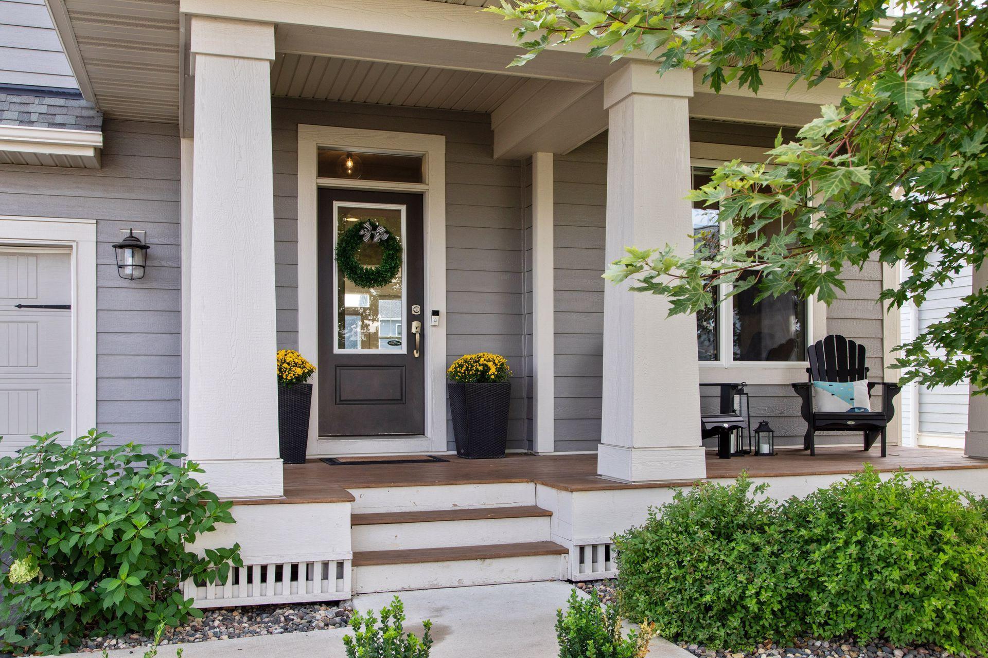 Covered front porch with white columns, a wreath on the dark door, yellow flowers, and a black chair.