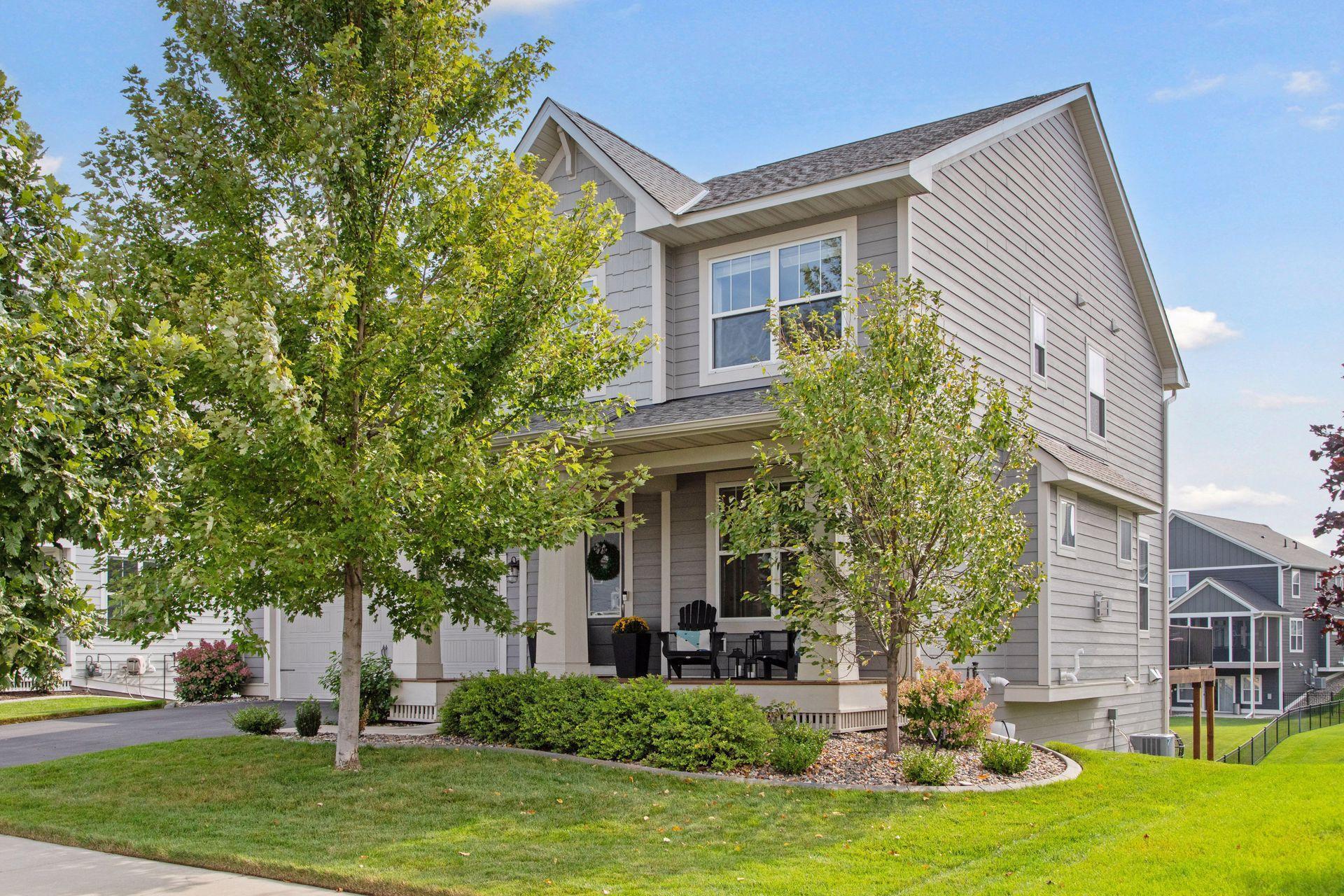 Two-story gray house with a covered porch, leafy trees, and landscaped front yard.