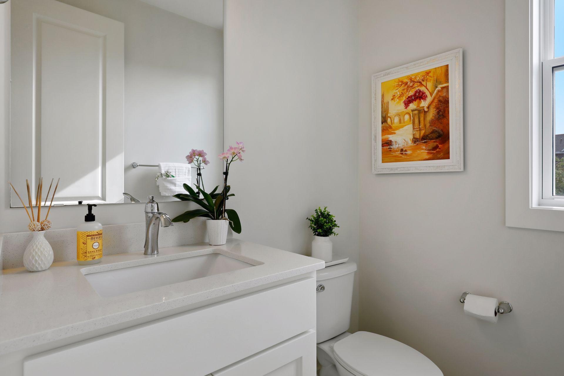 Bright bathroom with a white vanity, chrome faucet, pink orchids, and framed autumn artwork.