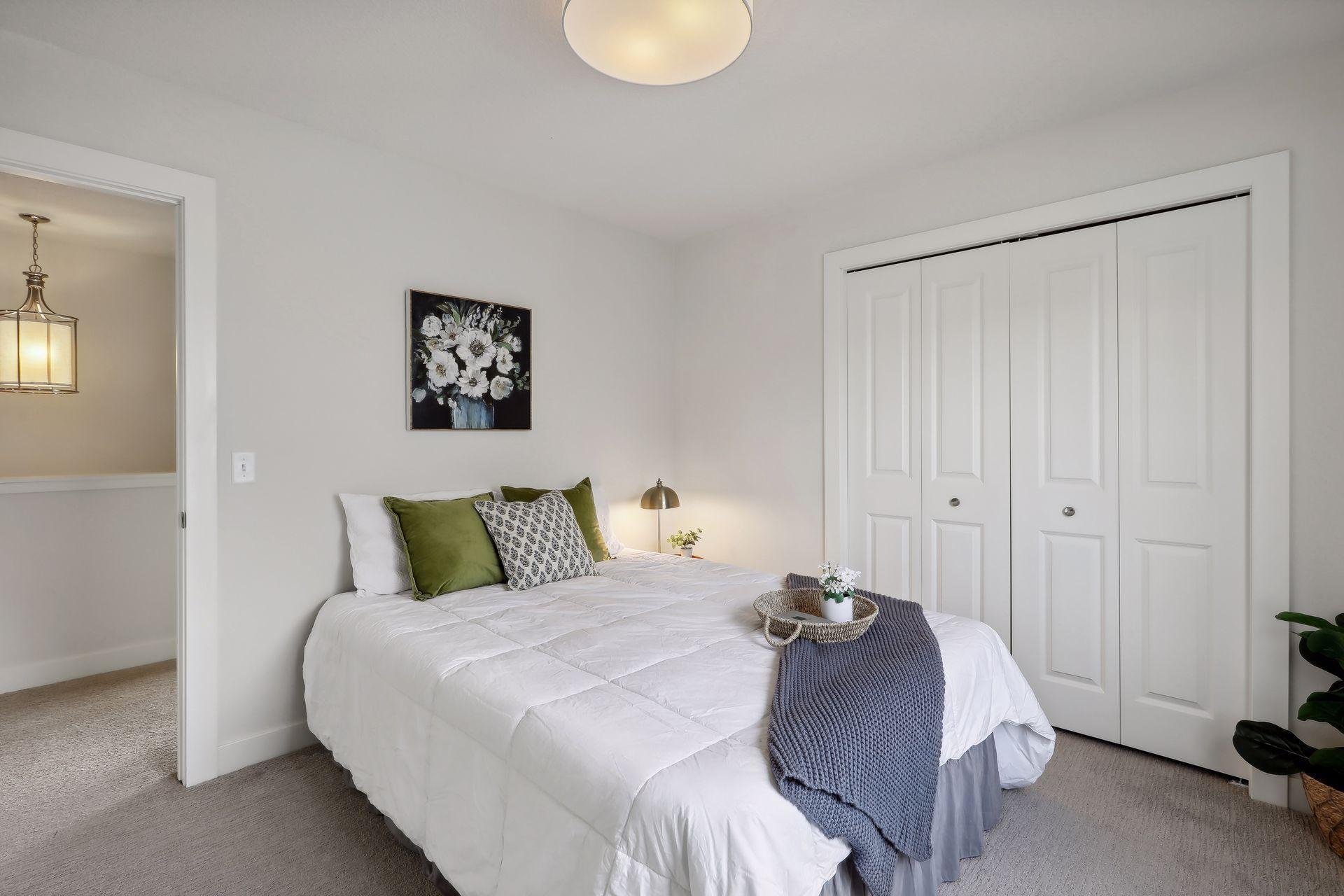 Staged bedroom with a white bed, green pillows, blue-gray throw, and white paneled closet doors.
