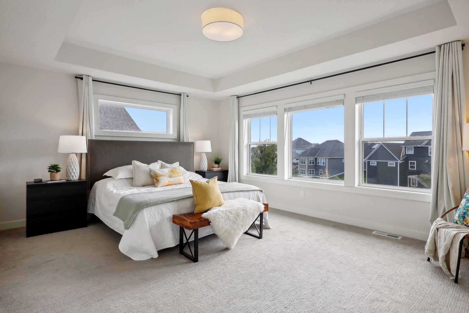 Staged bedroom with a large bed and wide windows overlooking neighboring homes.
