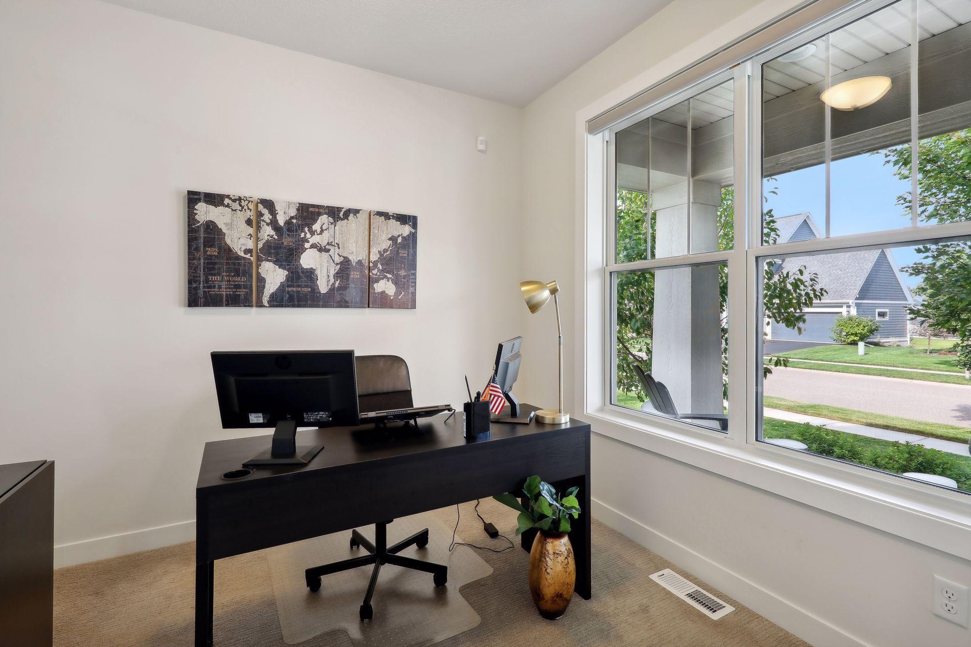 Furnished home office with a dark desk and large windows overlooking a residential street.