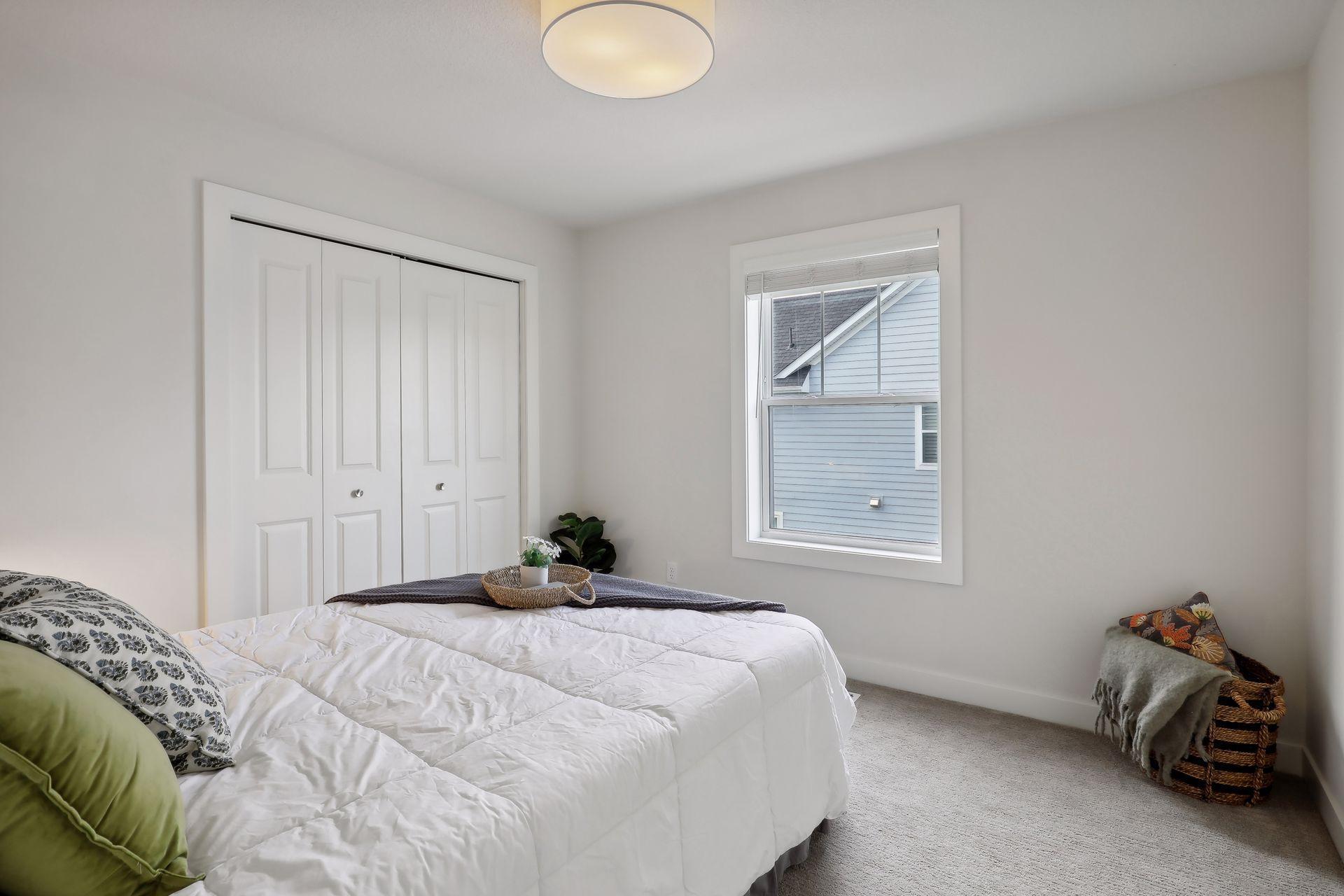Staged bedroom with a white bed, paneled closet doors, and a window with blinds.