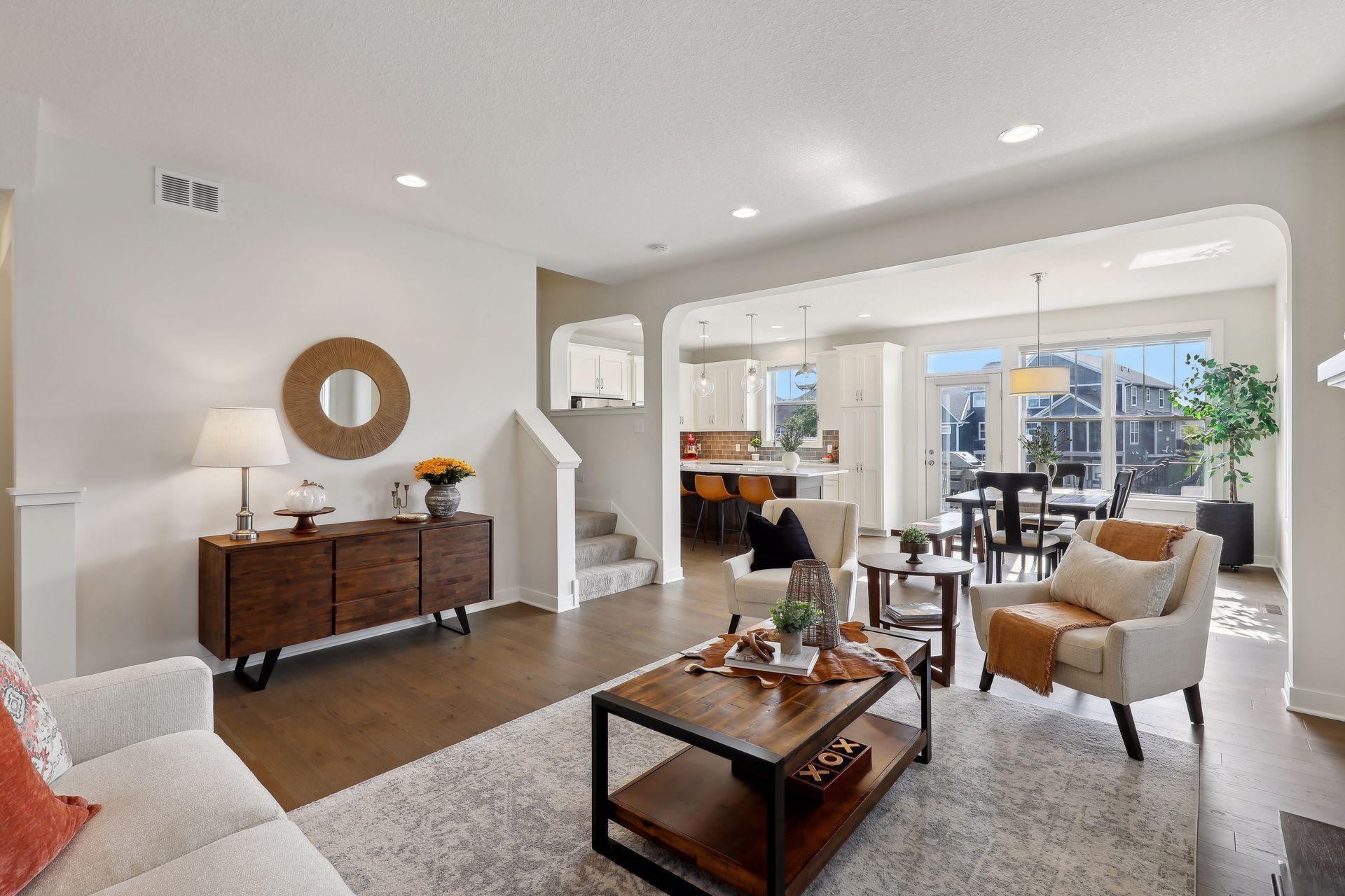 Staged living room opening to a dining area and white kitchen, with large windows and dark wood furnishings.