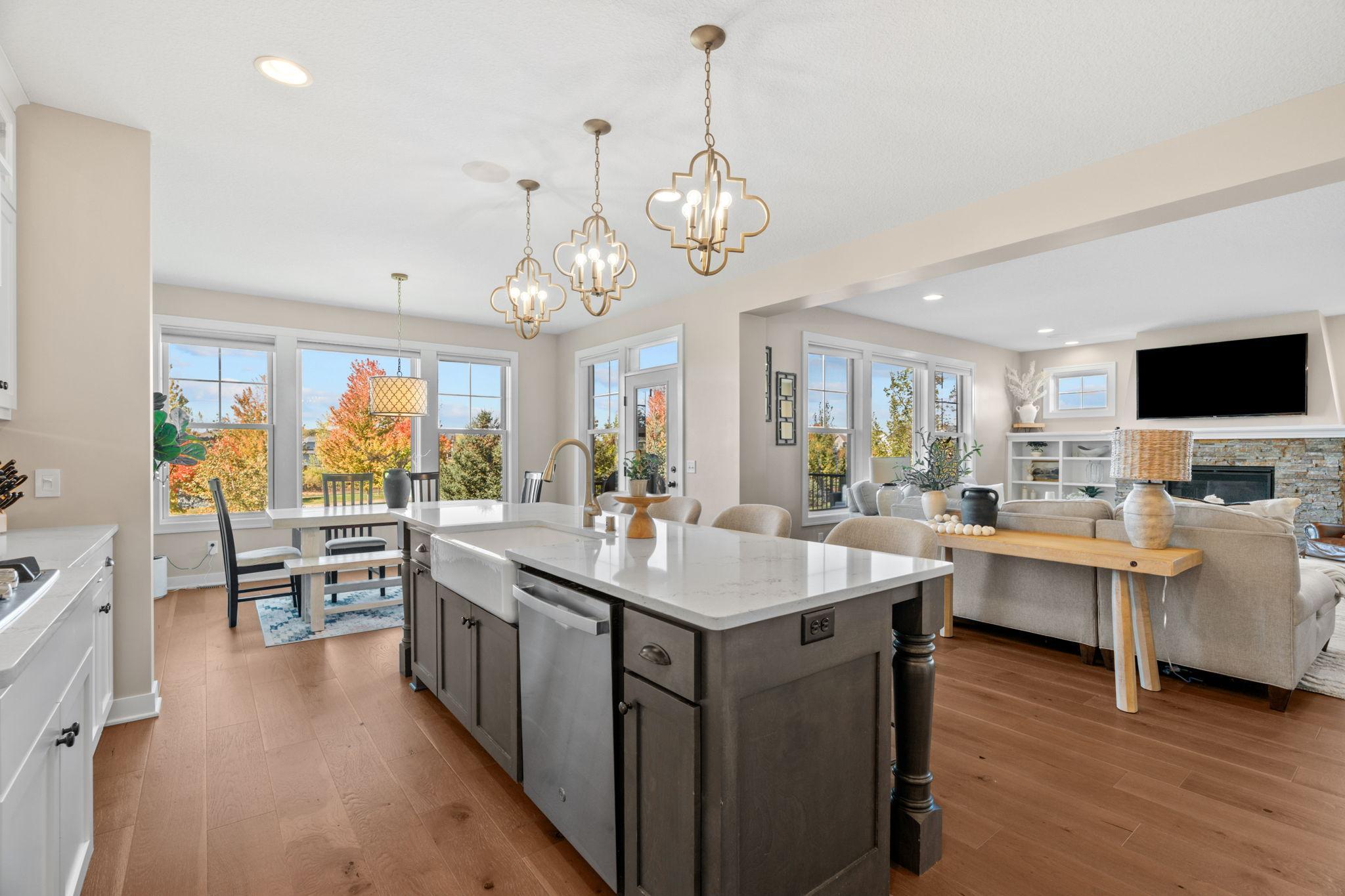 Open-plan kitchen and living room with a large island, gold pendant lights, and autumn trees visible through the windows.