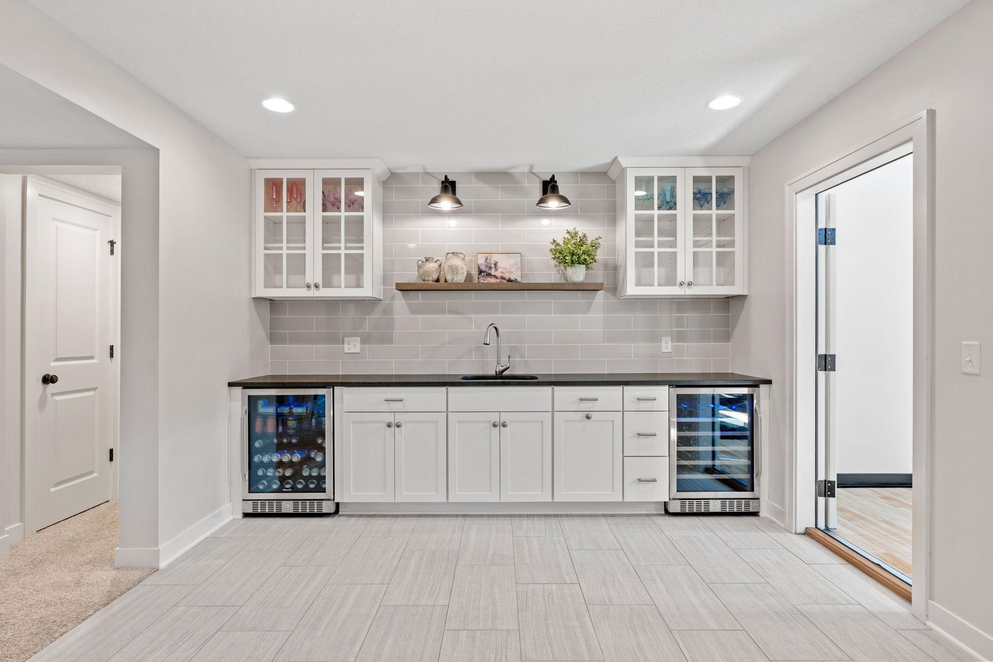 Basement wet bar with white cabinets, gray tile backsplash, sink, and two glass-front beverage refrigerators.