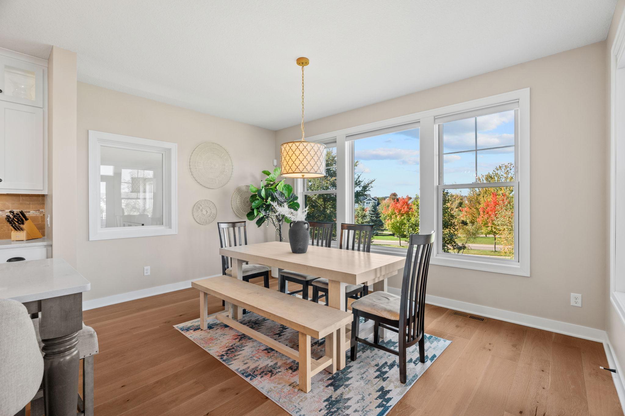 Dining room with a light wood table and bench beside large windows overlooking autumn trees.