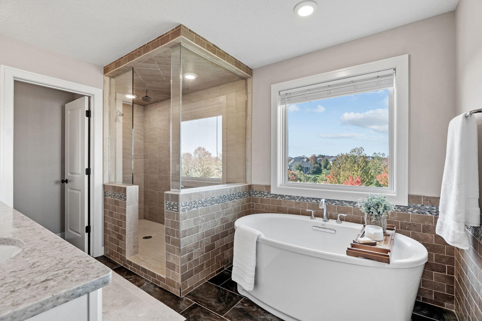 Bathroom with a glass-enclosed tiled shower and freestanding tub beside a window overlooking autumn trees.