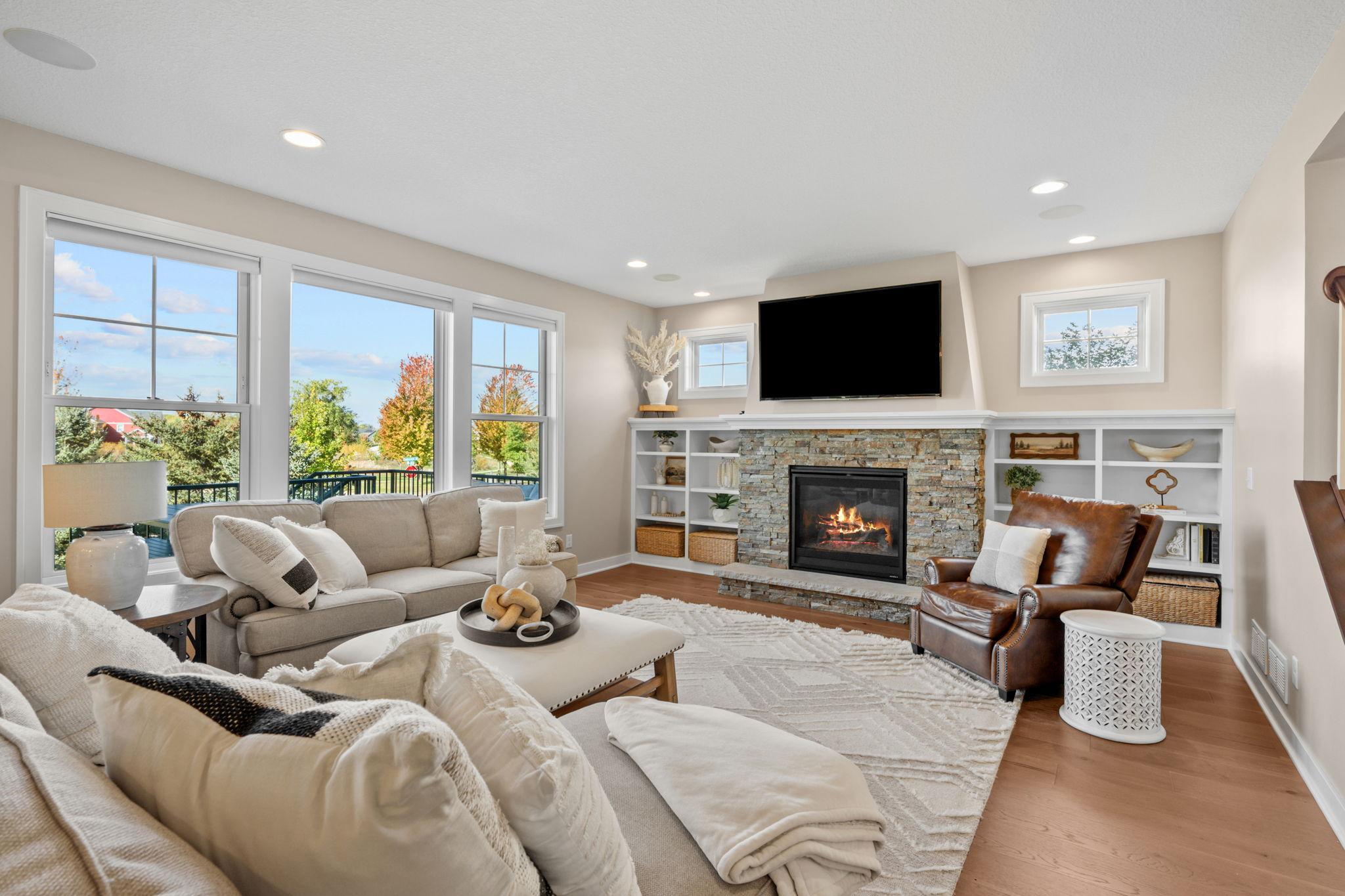 Furnished living room with a stone fireplace, built-in shelves, and large windows overlooking autumn trees.