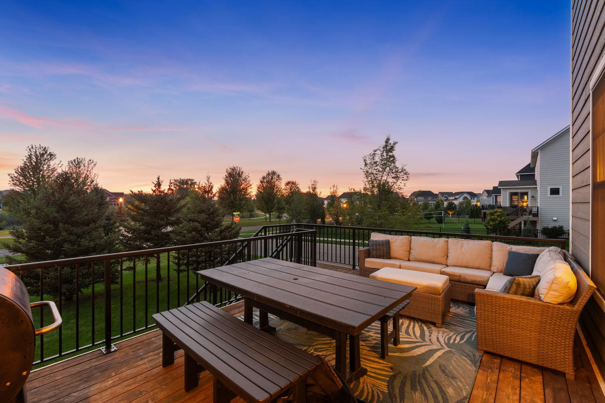 Furnished deck with wicker seating and dining table overlooking a neighborhood at sunset.