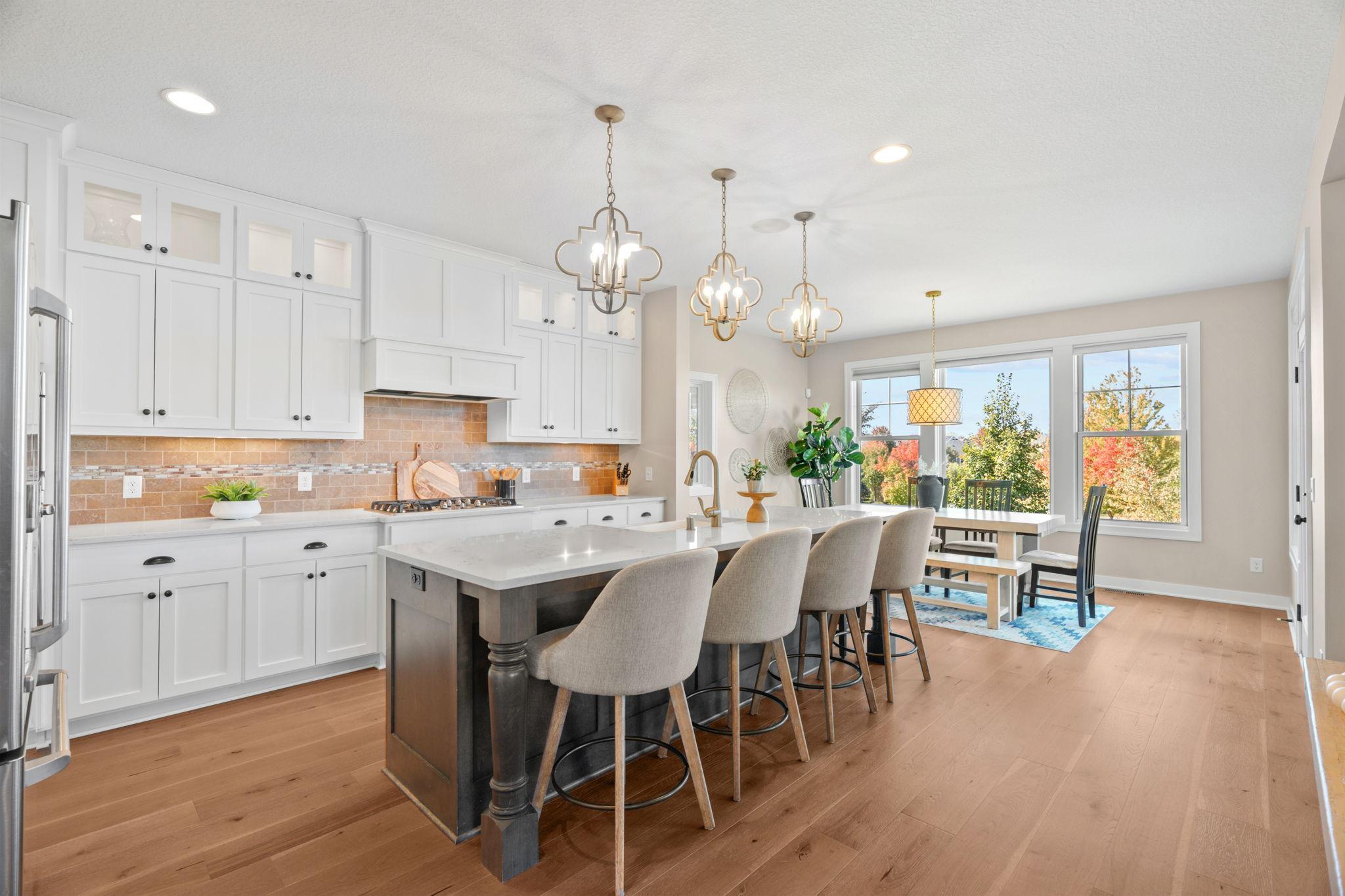 Bright kitchen with white cabinetry, a large island, wood floors, and windows overlooking autumn trees.