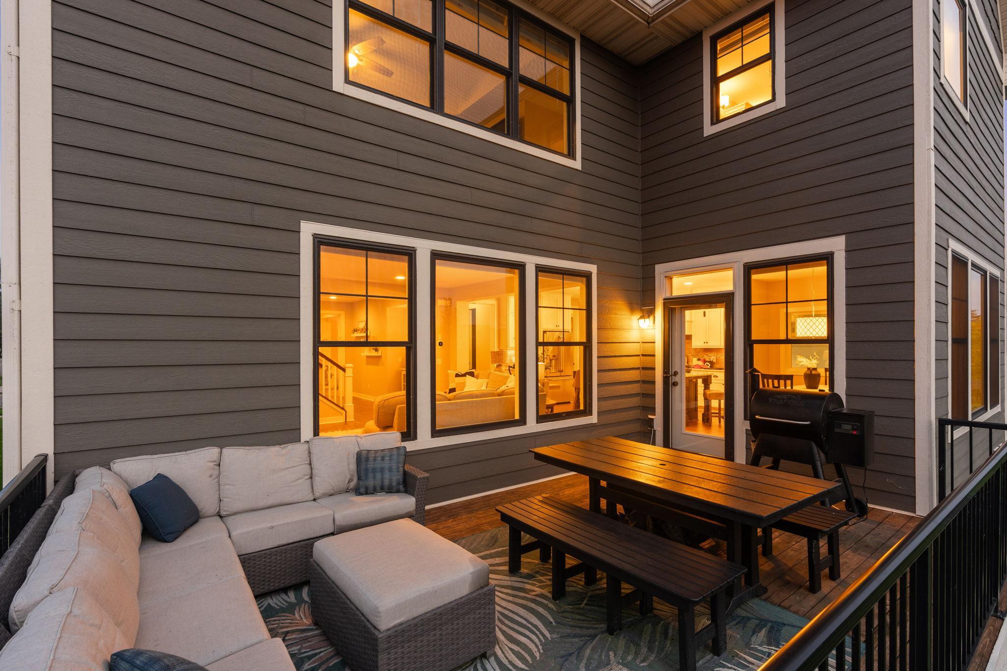 Furnished deck with sectional seating, wooden dining table, and grill beside a warmly lit home.