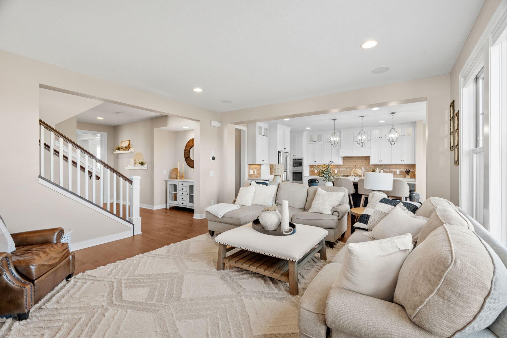Open-concept living room with neutral sectional sofas, a staircase, and a bright white kitchen beyond.