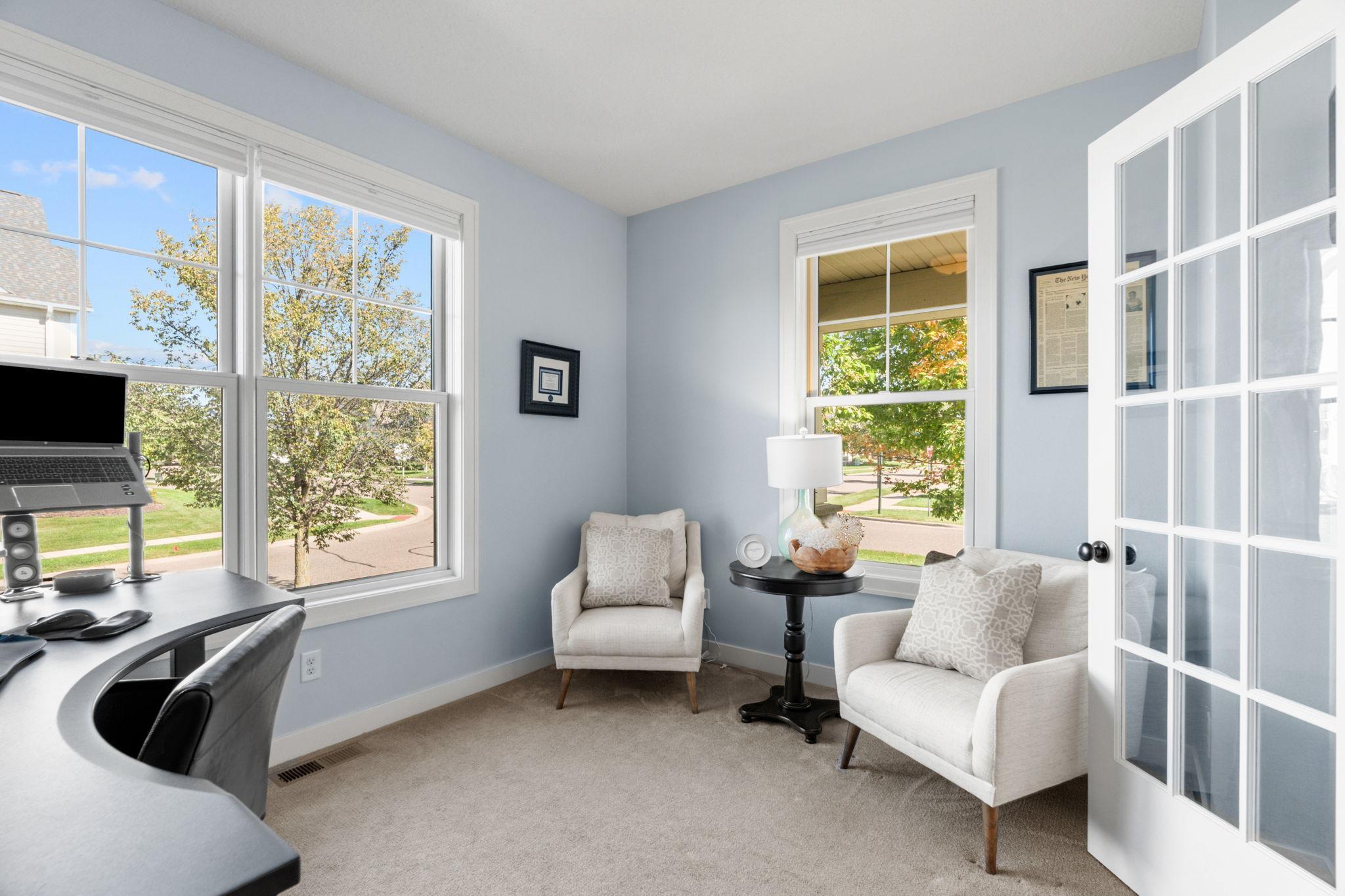 Home office with a curved desk, two white armchairs, and large street-facing windows.