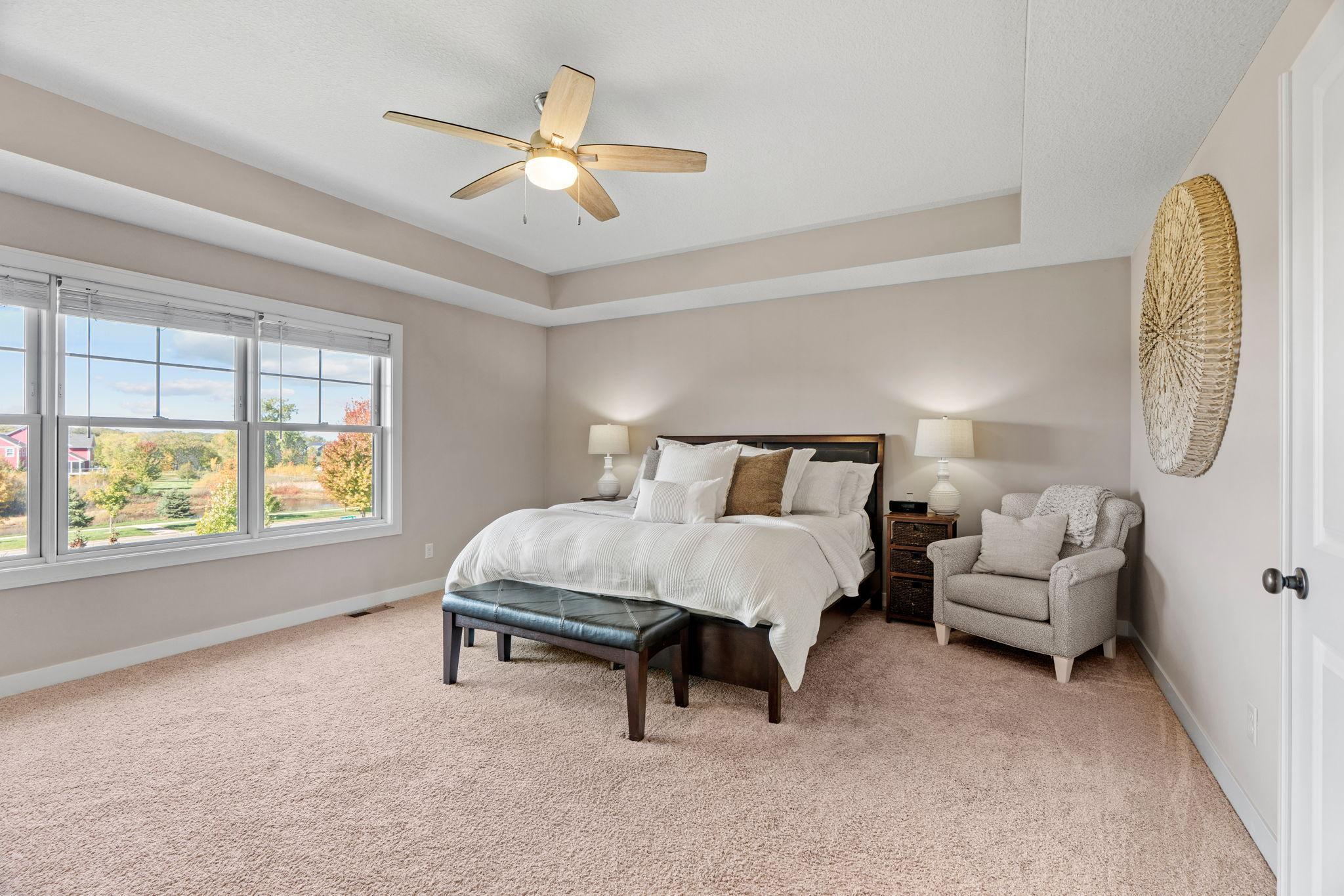 Furnished bedroom with a white bed, tray ceiling, and large windows overlooking autumn trees.