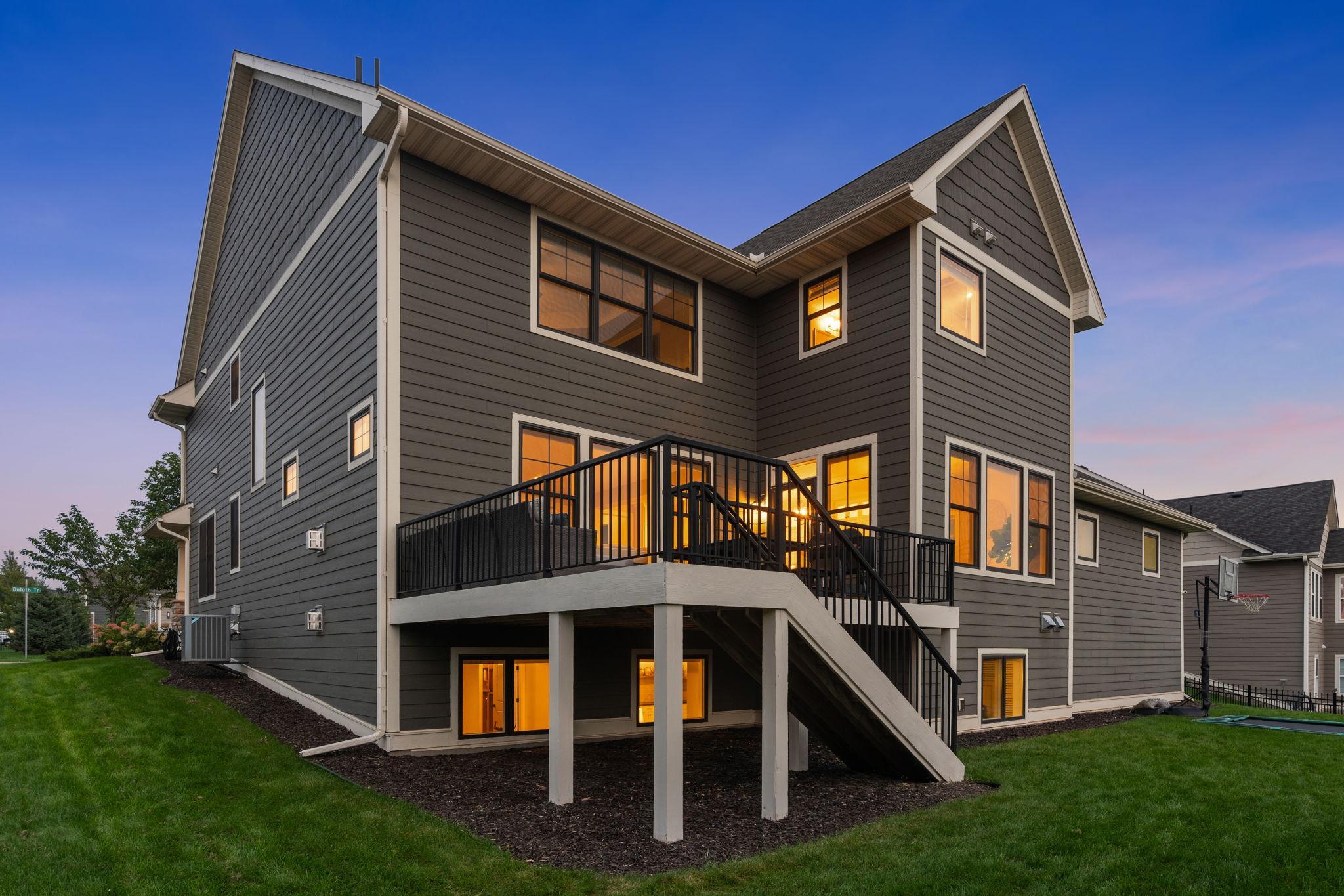 Two-story gray house at dusk with a raised deck, black railings, and stairs to the lawn.