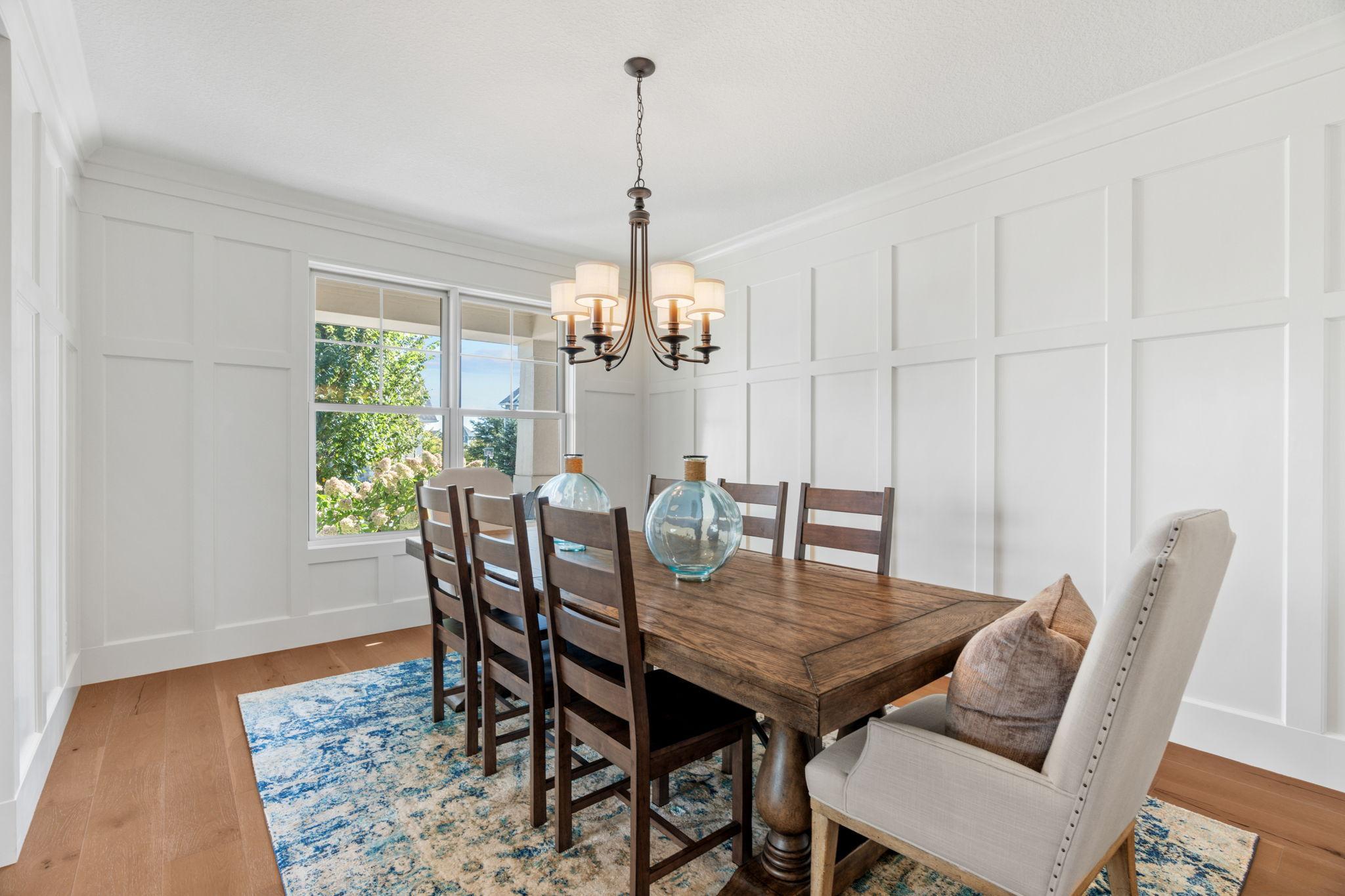Dining room with a dark wood table, white paneled walls, and large windows overlooking greenery.