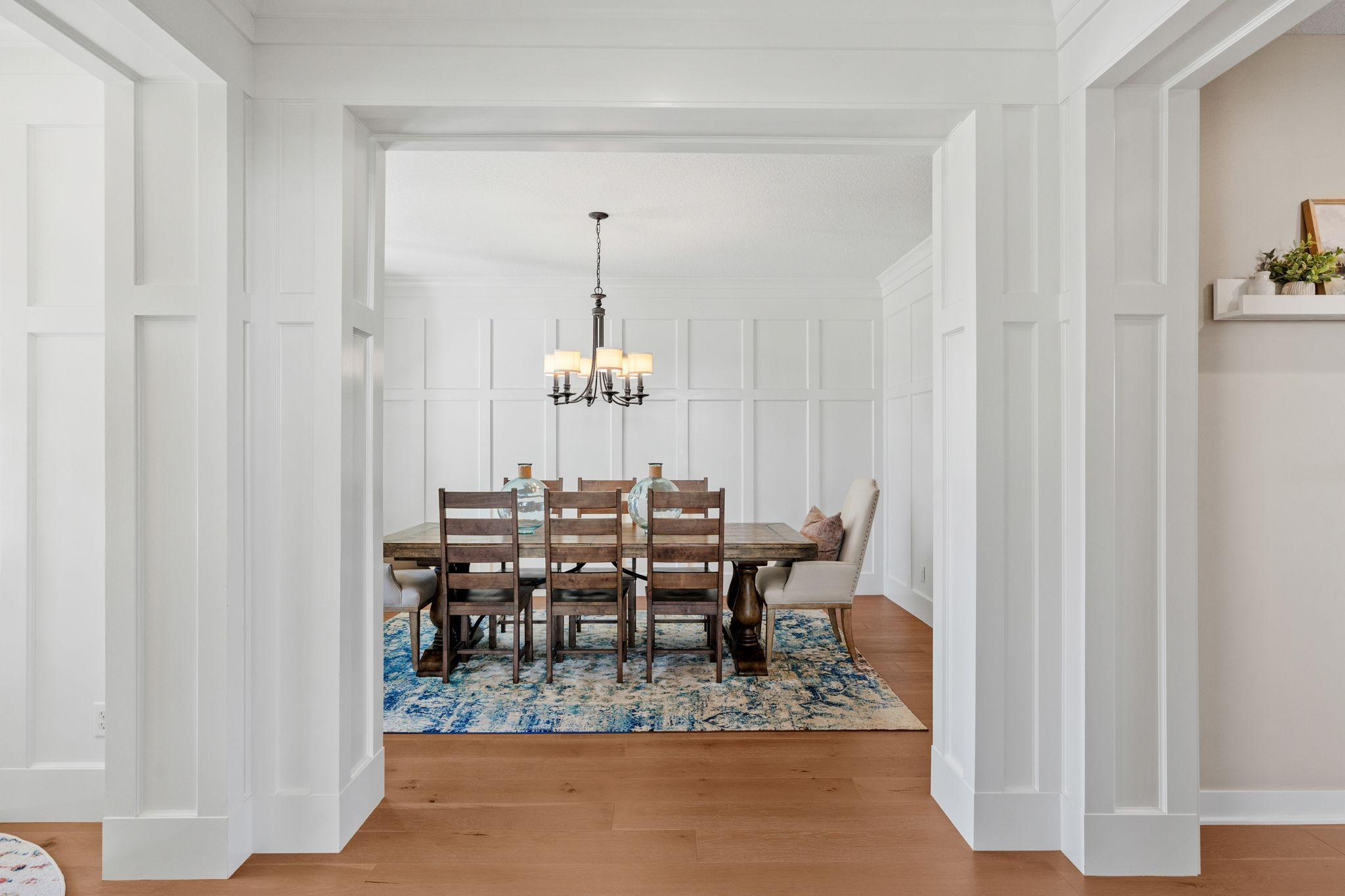 Dining room with white paneled walls, a wood table, chandelier, and blue patterned rug.