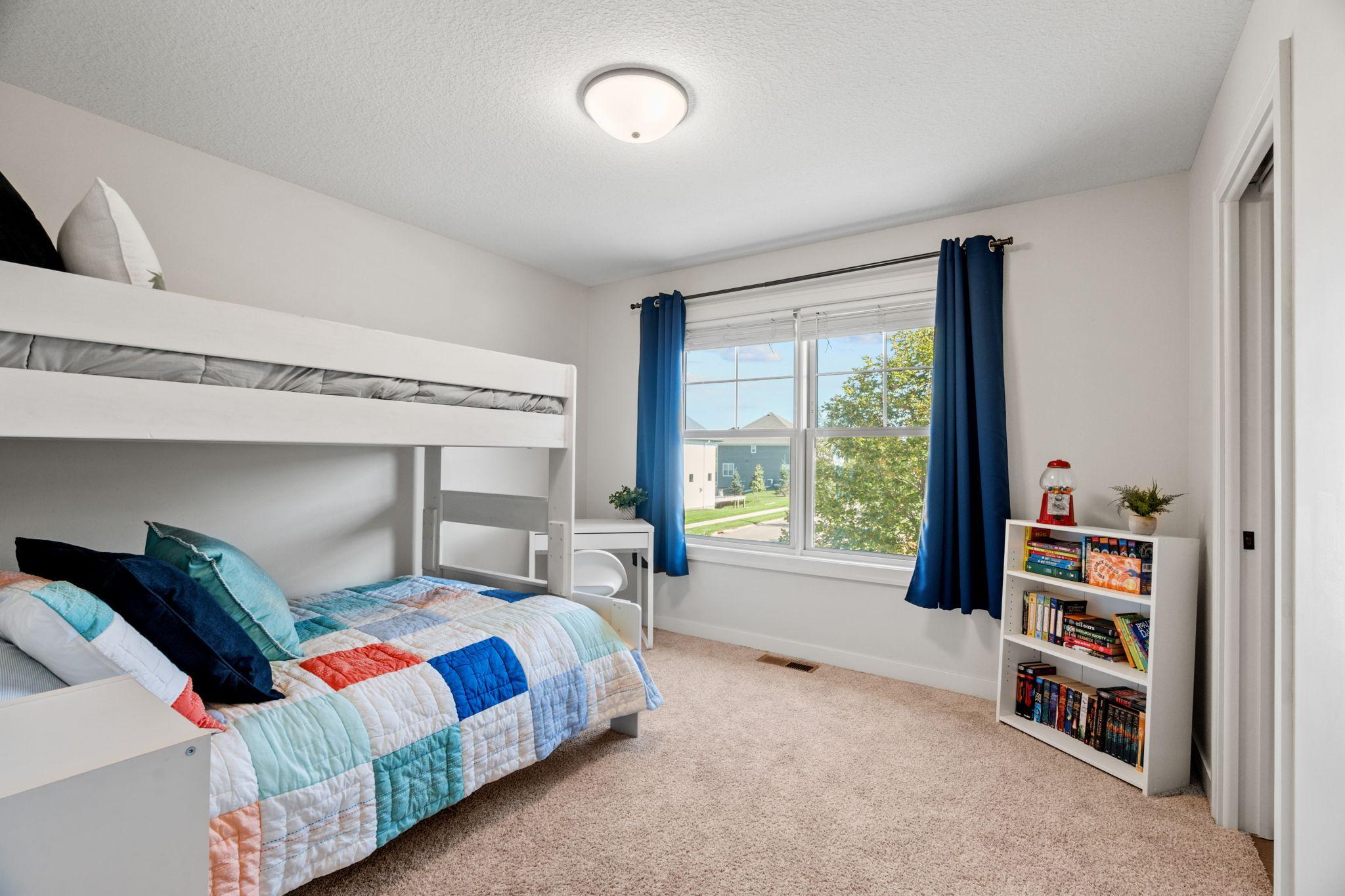 Bedroom with a white bunk bed, colorful quilt, blue curtains, and a bookcase of books and games.