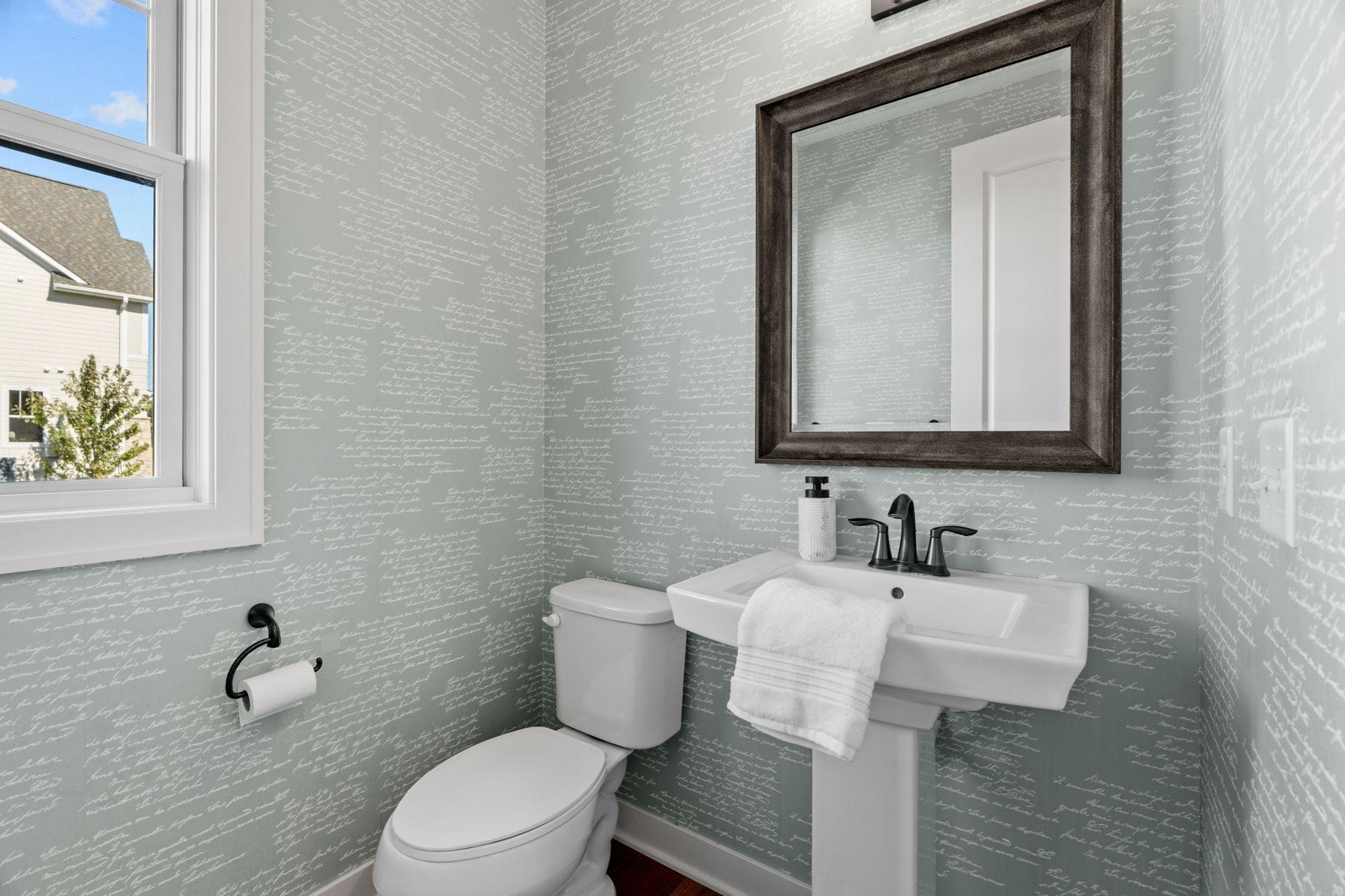 Powder room with script-patterned wallpaper, pedestal sink, toilet, and dark-framed mirror beside a window.