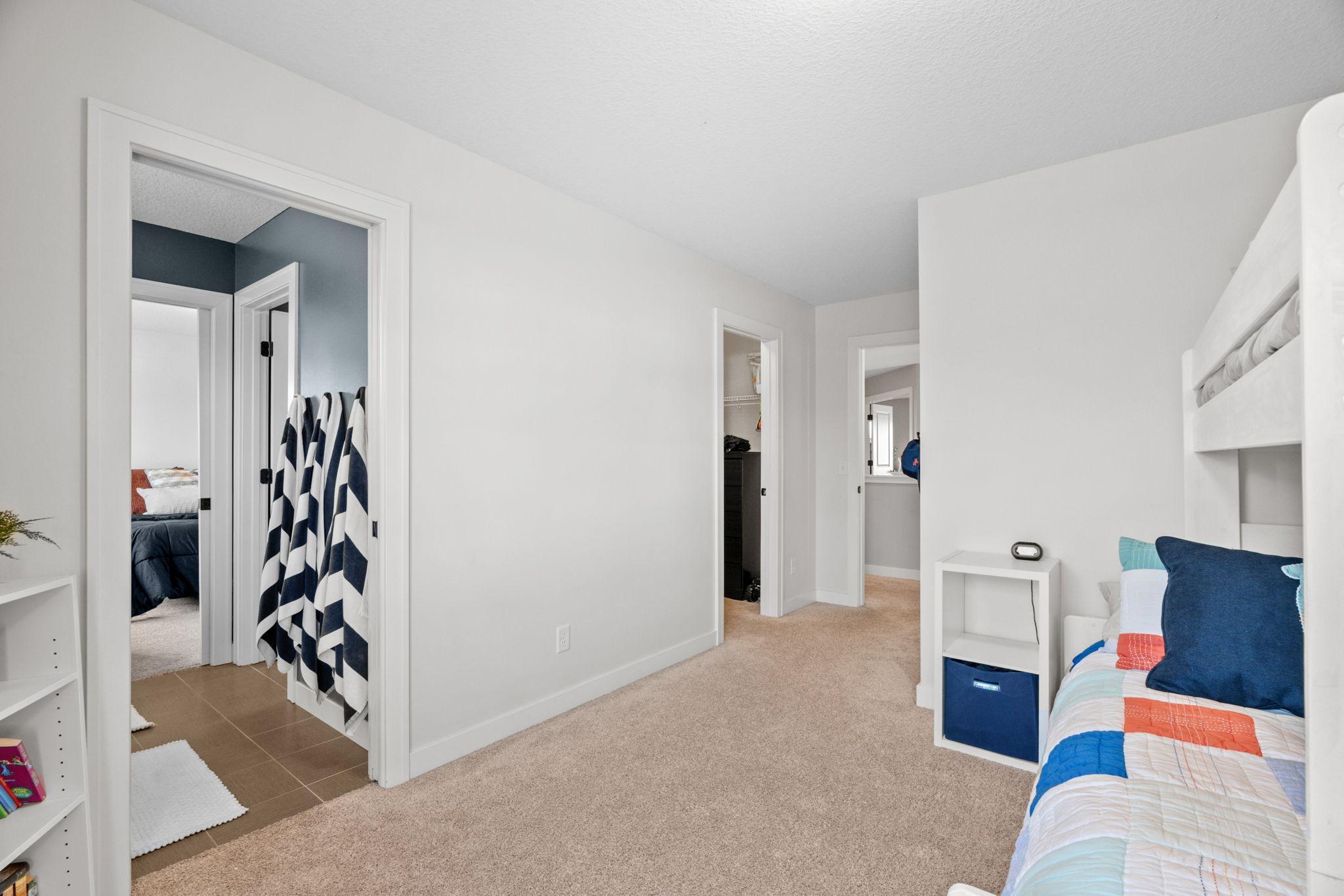 Bedroom with a white bunk bed, colorful quilt, beige carpet, and open doorways to adjoining rooms.