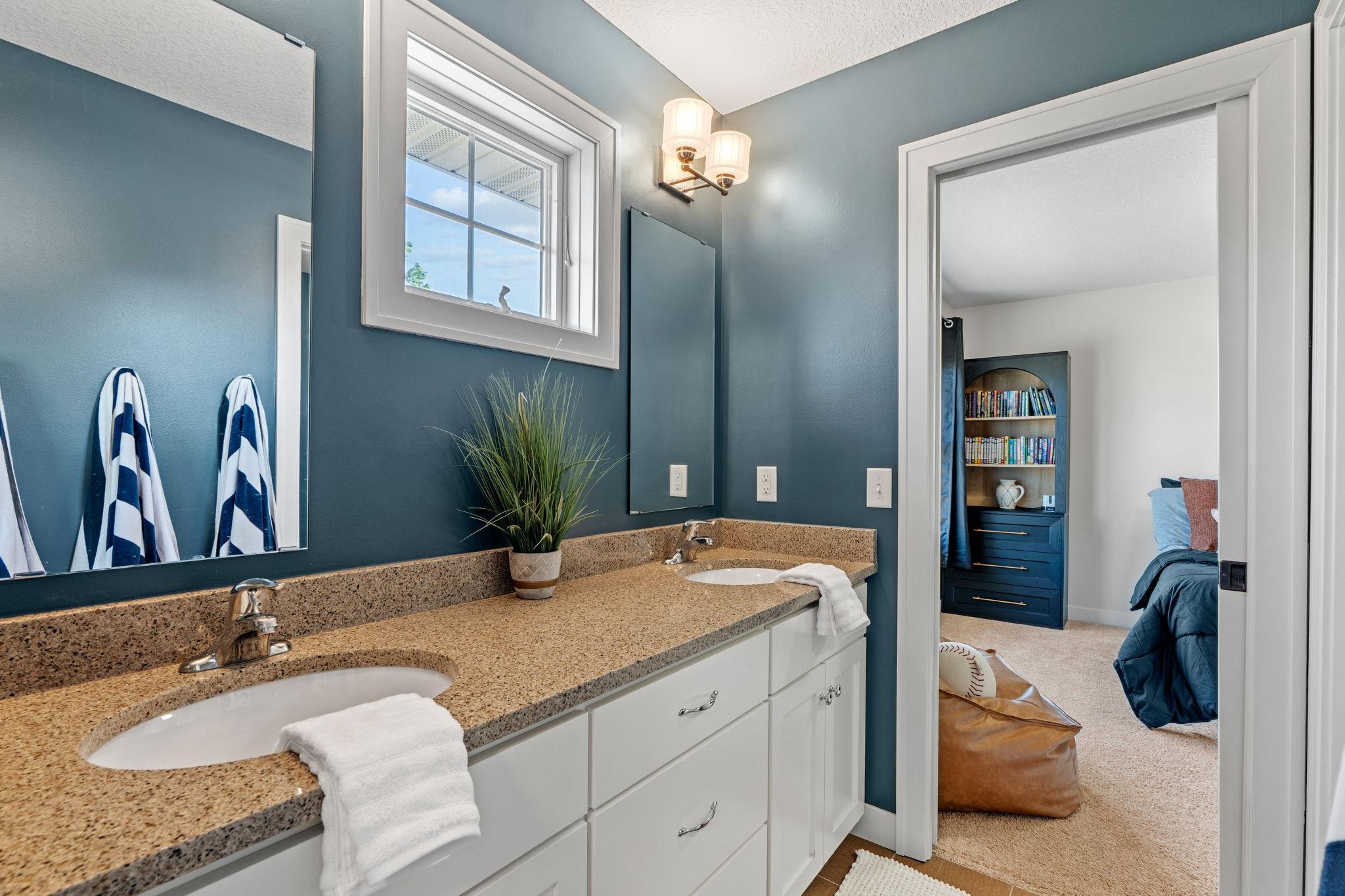 Bathroom with a double-sink granite vanity and blue walls, opening into a bedroom.