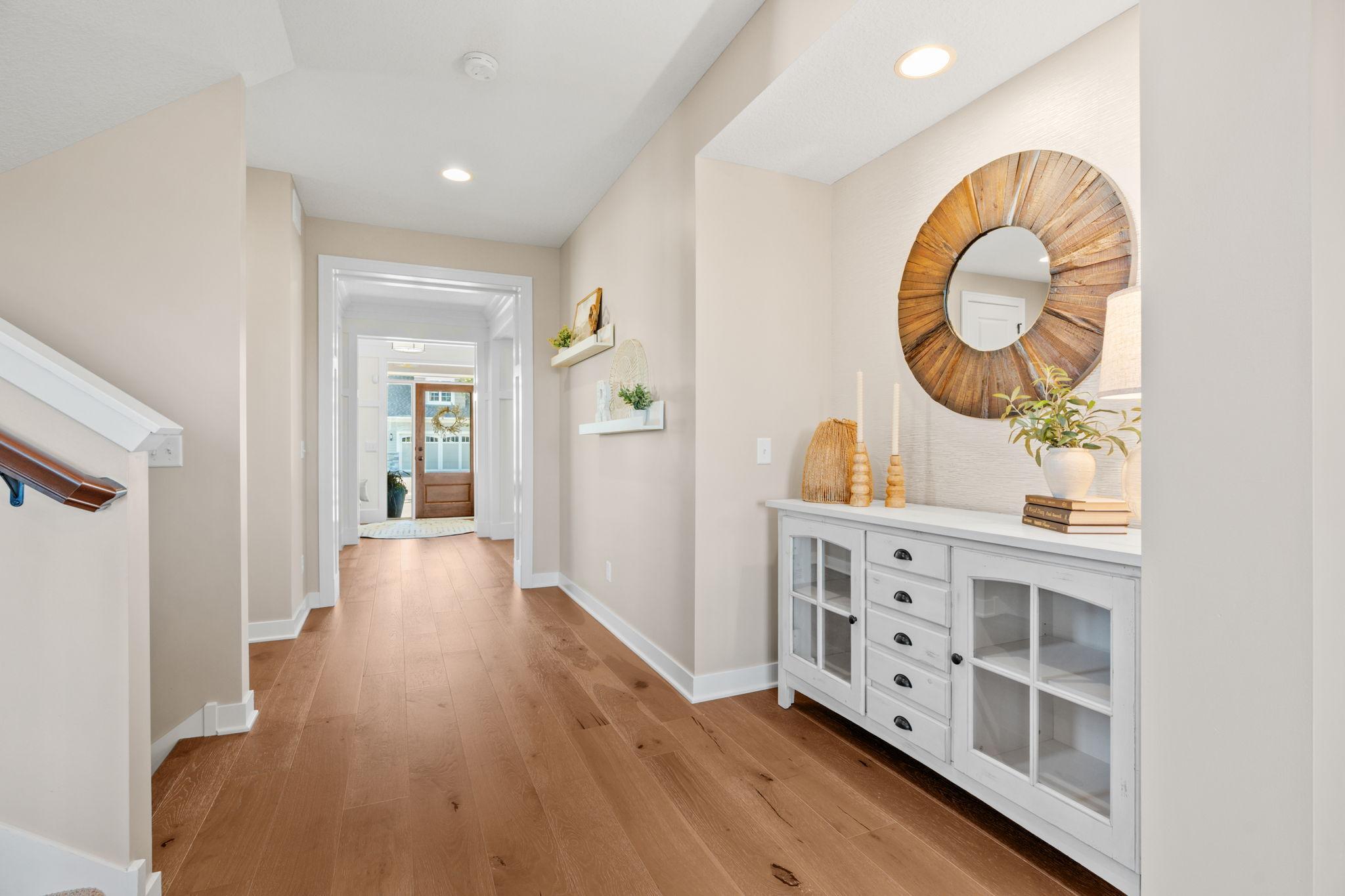Staged entry hall with warm wood floors, a round wood-framed mirror, and white console cabinet.