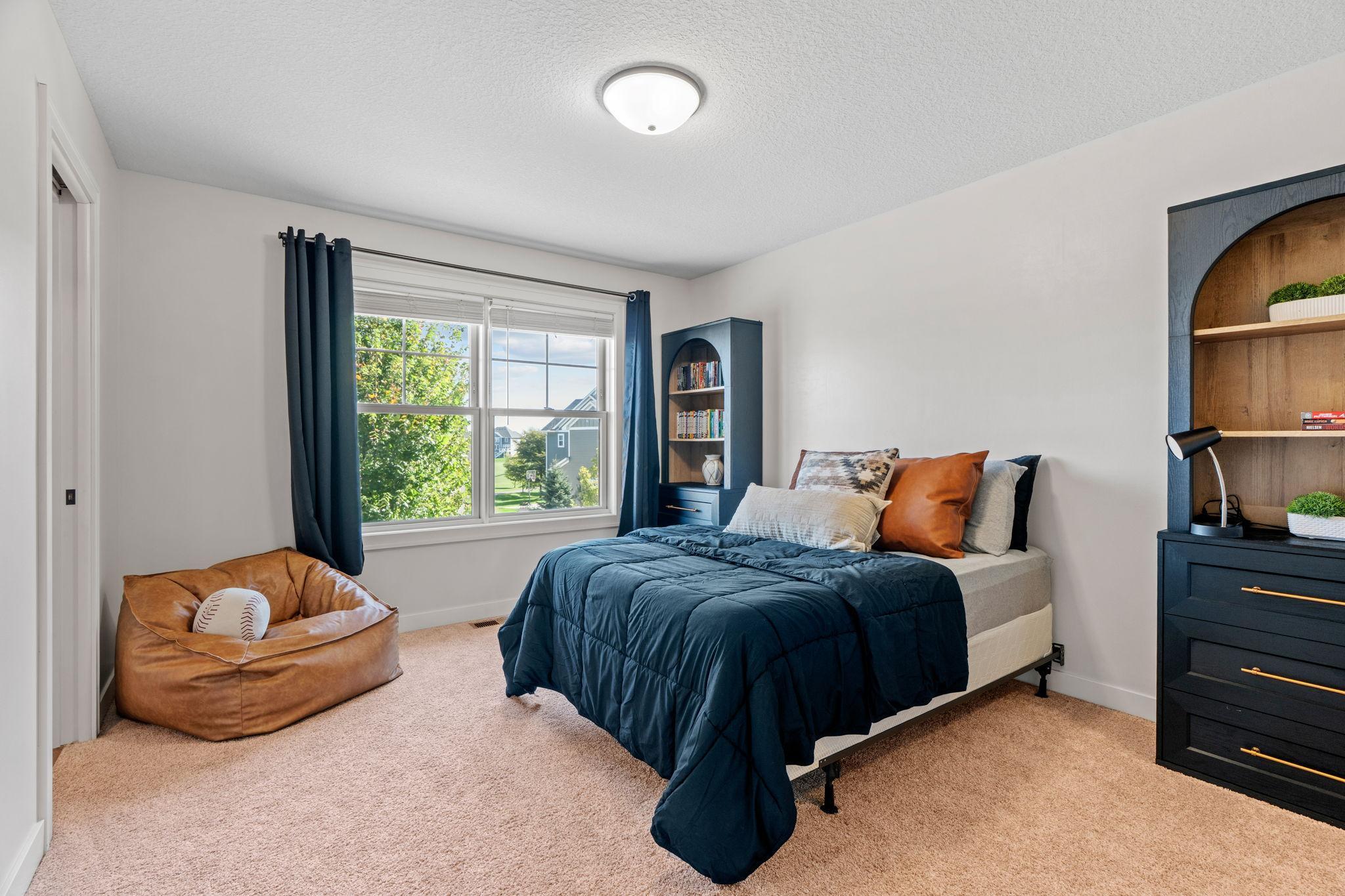 Furnished bedroom with a navy-quilted bed, blue curtains, and a tan beanbag holding a baseball.