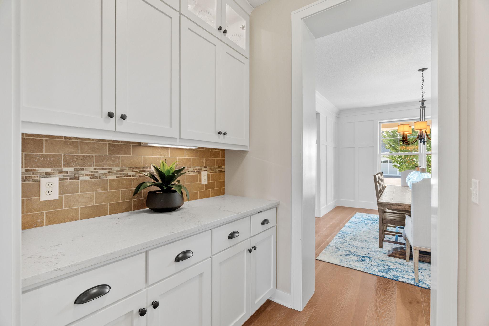 White kitchen with tan tile backsplash opens to a dining room with a wood table and blue rug.