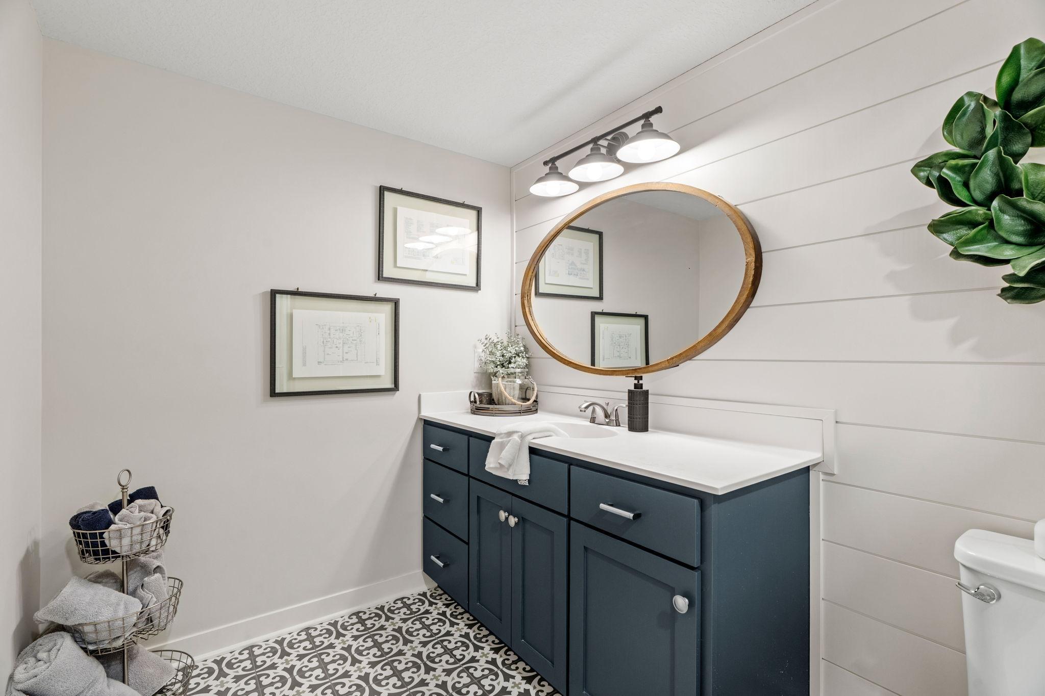 Bathroom with a dark blue vanity, round wood-framed mirror, shiplap walls, and patterned tile floor.